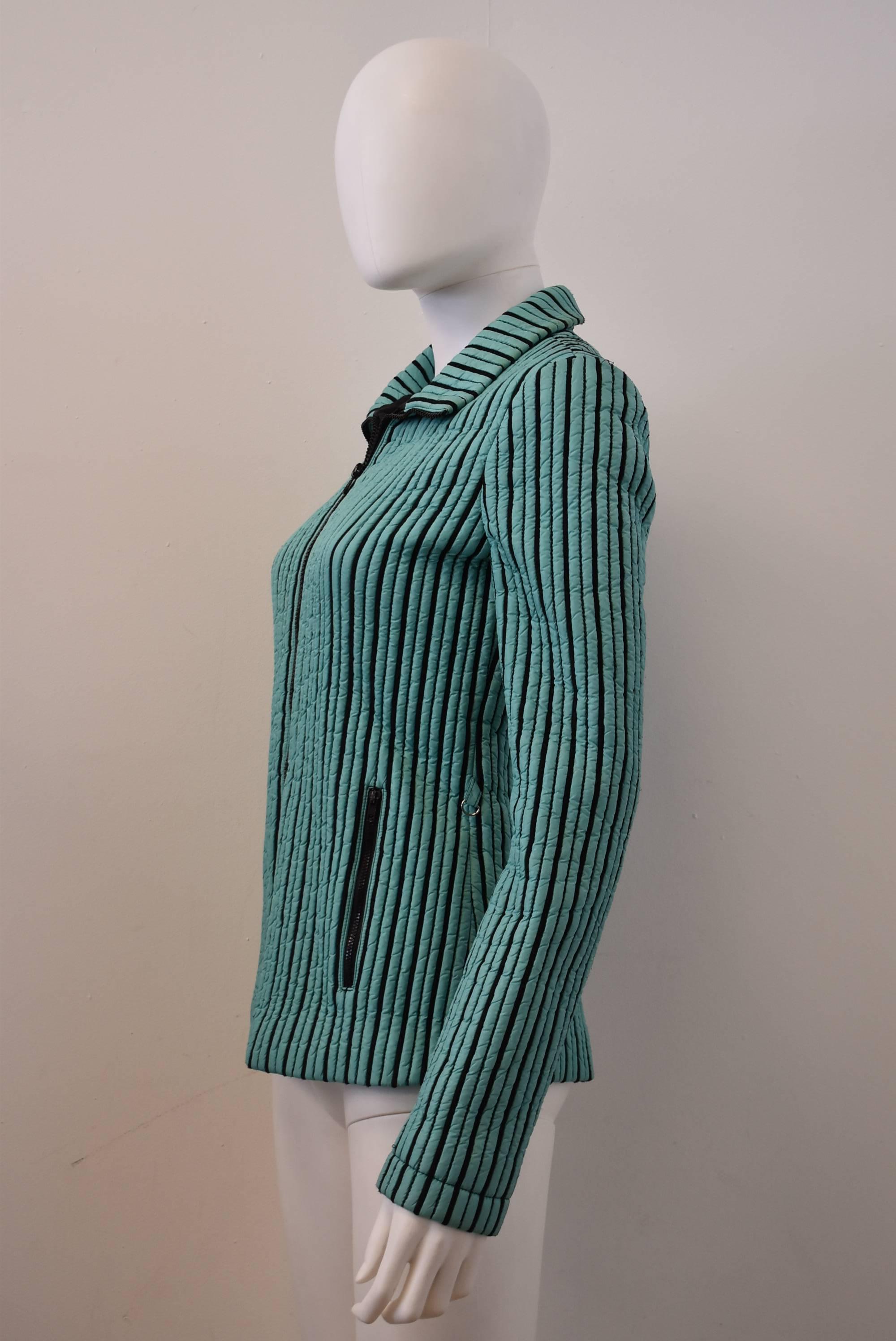 A green turquoise and black ‘striped’ jacket created using piping and stitching techniques. The jacket has a simple, short shape with a zip front fastening and two zip-close pockets at the hip. The jacket is made from a sporty synthetic material