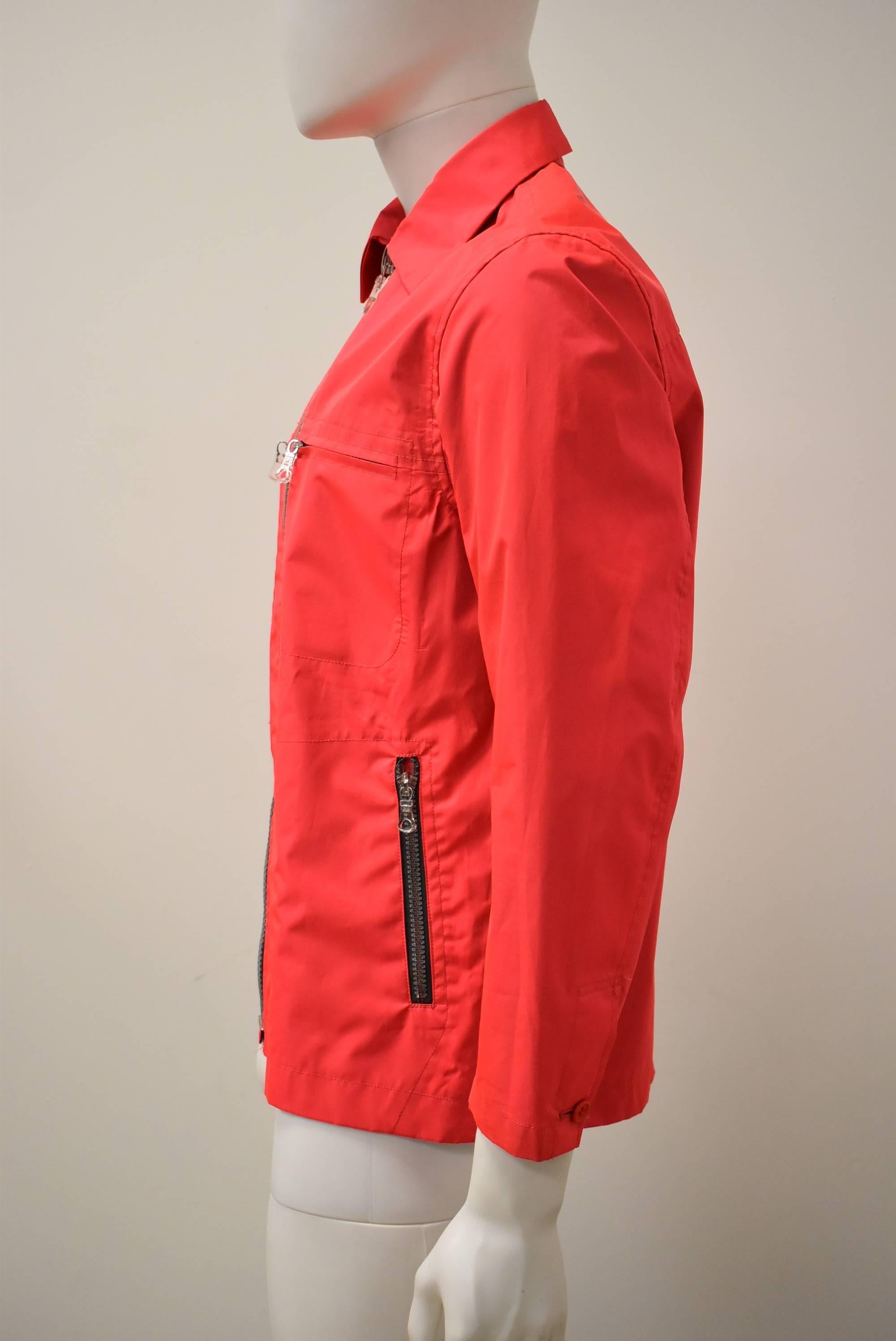 A bright neon red lightweight sports jacket from Issey Miyake’s mainline collection. The jacket has a simple, straight, short shape, falling to just below the waist. There is an oversize clear zip-fastening detail on the front. There are also two