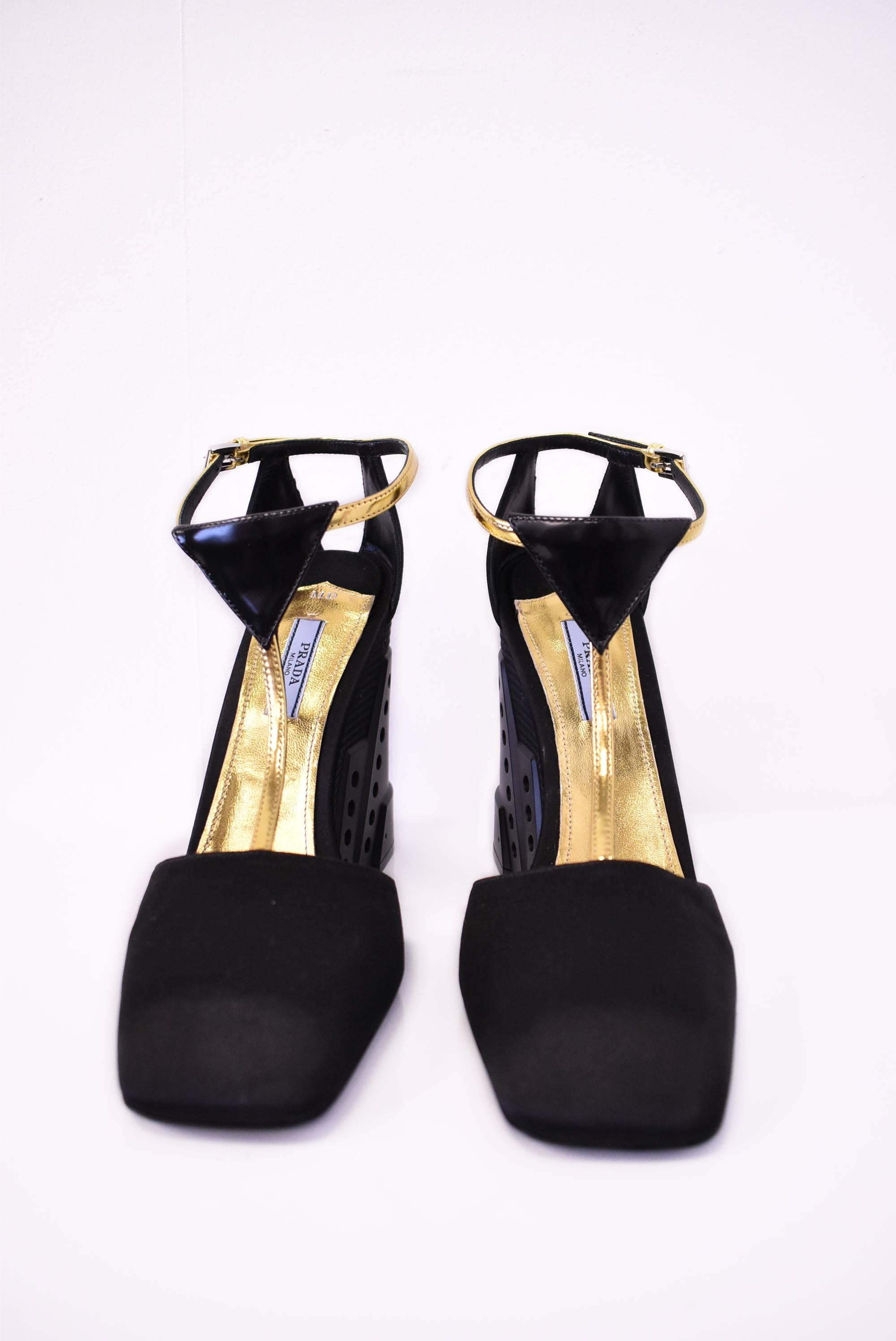 A pair of unworn and brand new black Prada wedge heels. The shoes are made from black silk satin with a closed square toe and gold T-strap ankle fastening. The wedge heel has a 5” height and a futuristic architectural, faux metal, perforated design.