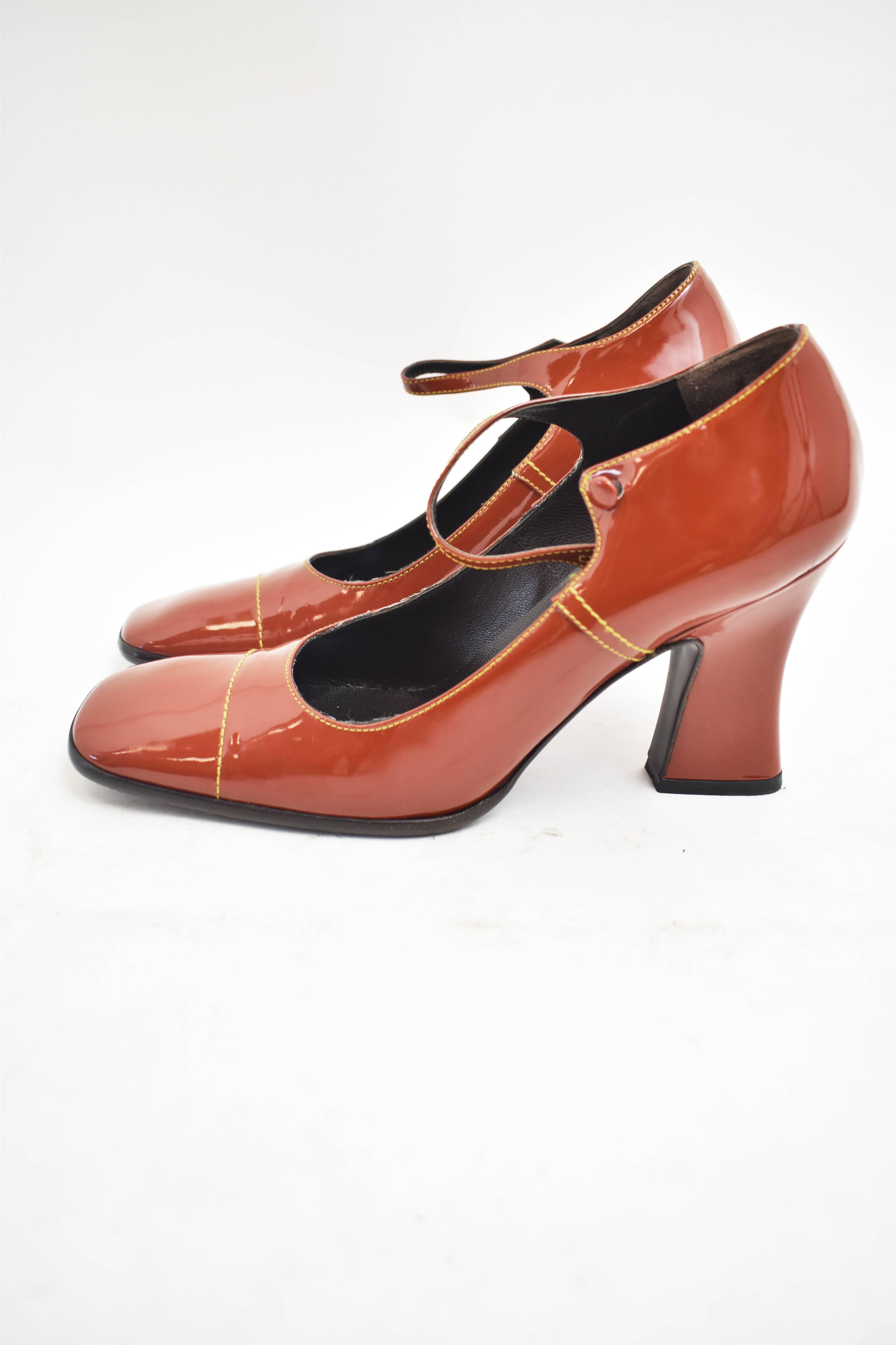 A pair of classic Prada Mary Jane Shoes made from a rust brown patent leather. The shoes have a slim shape, square toe, contrast color stitching and a strap across the ankle. The heel is a medium height with a chunky shape and a curved silhouette.