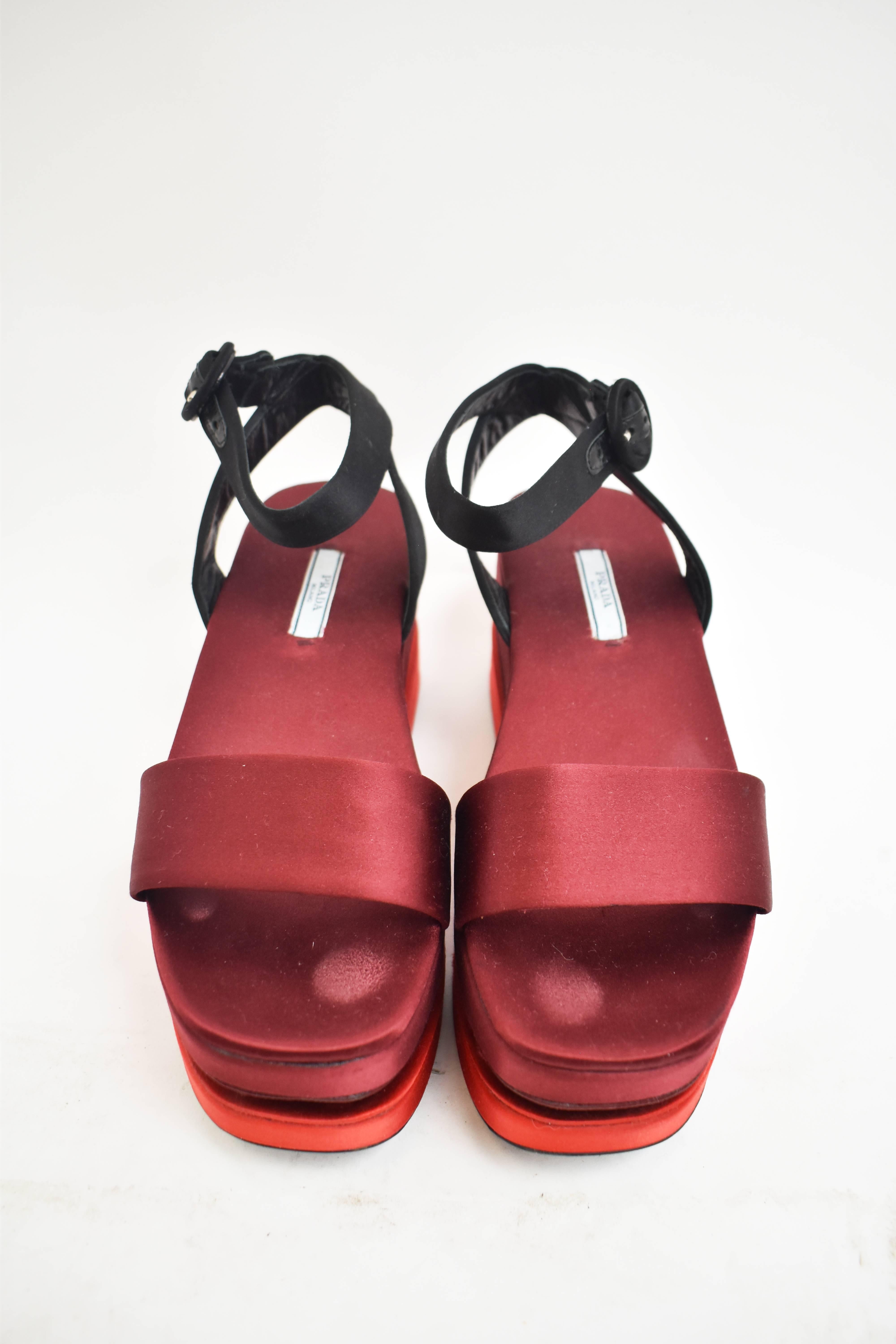 A pair of Prada sandals made from a dark red satin with contrast lighter red satin sole. The sandals have an interesting platform heel that is made from black lacquer wooden blocks that create the effect of a ‘floating’ sole. They have a single