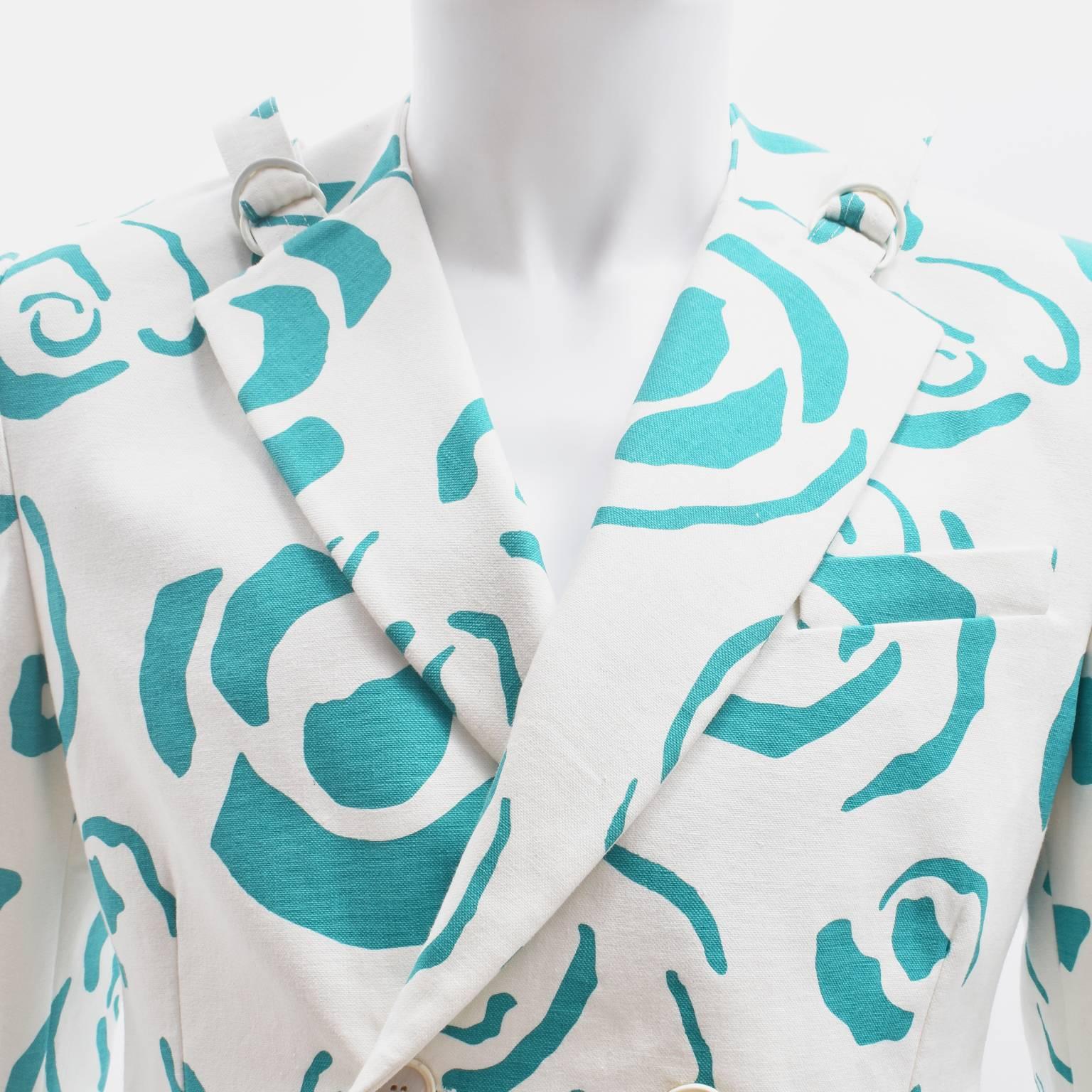 A white double breasted jacket with an all-over turquoise green rose print by British designer J.W. Anderson. The jacket has a classic double-breasted design with four buttons, two pockets at the hip and a boxy silhouette. However, the collar