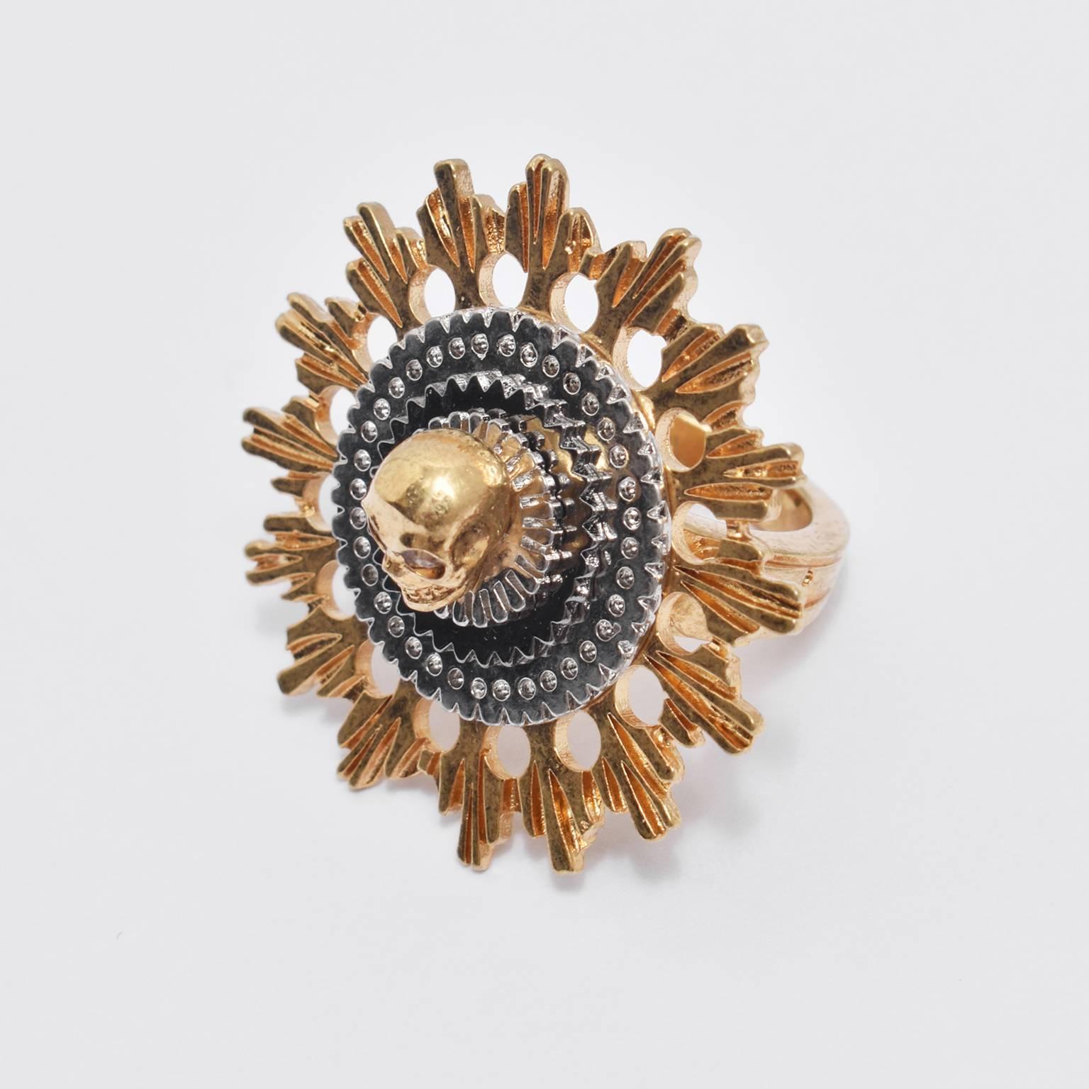 A stunning gold plated ring from Alexander McQueen. The ring features McQueen’s signature 3D skull design in a contrast silver roundel with a gold abstract, mechanical sunburst around the outside. The circular shape is attached to a thick gold band.