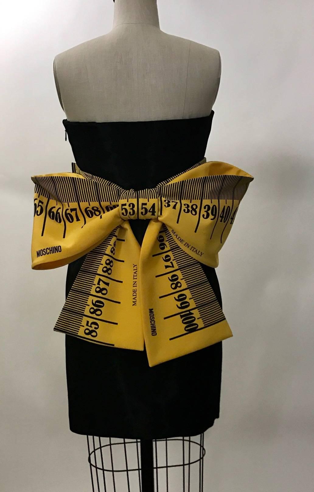 Recent Moschino Couture by Jeremy Scott black strapless dress with sweetheart neckline and wide measuring tape sash tied in a giant bow at back. Black material has a light sheen. Measuring tape reads 'Moschino' and 'Made in Italy.'  Built in corset