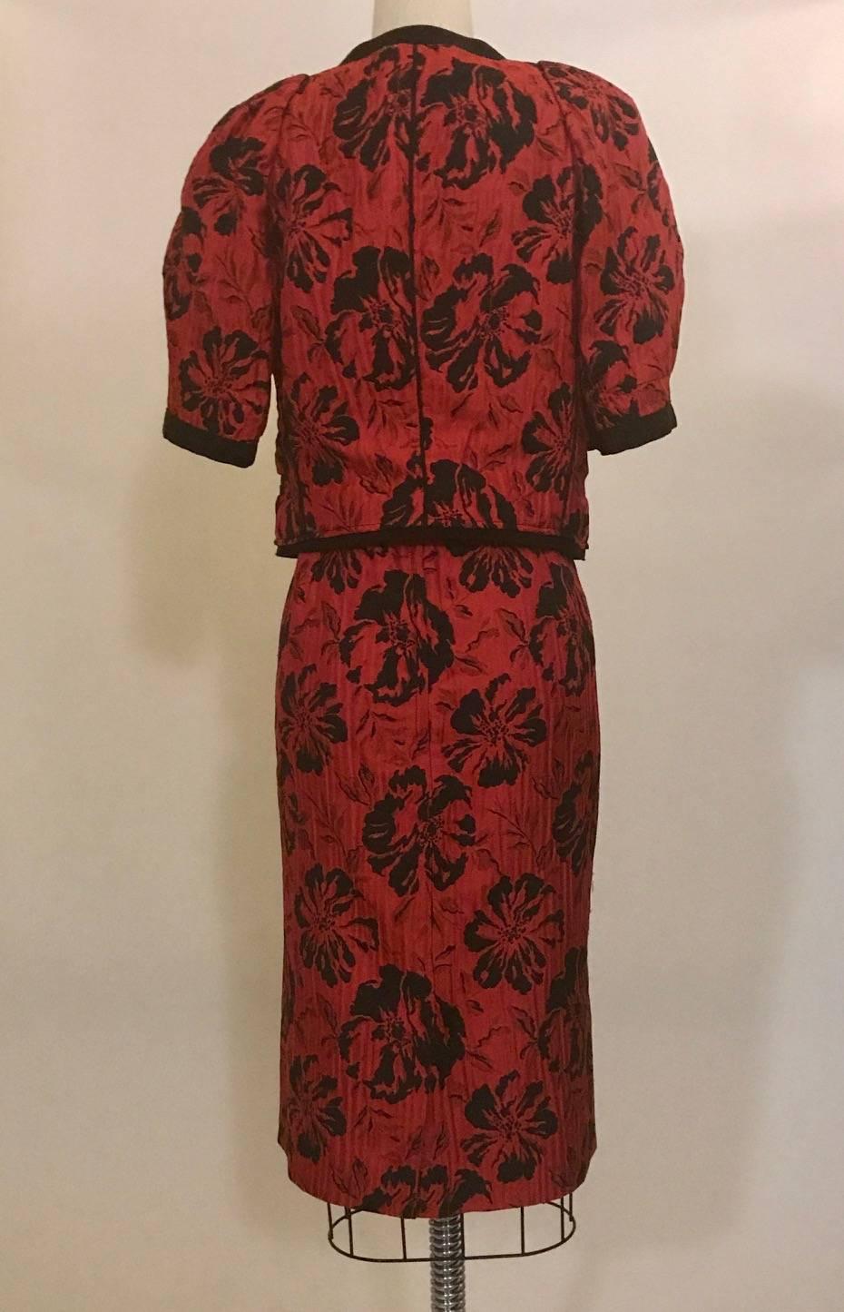 Dolce & Gabbana red and black jacquard skirt suit in a floral hibiscus print. Short sleeves, mock welt pockets at jacket, grosgrain trim. Jacket fastens at front with concealed snaps, skirt has back zip.

45% acetate, 33% silk, 22% polyamide.
Fully