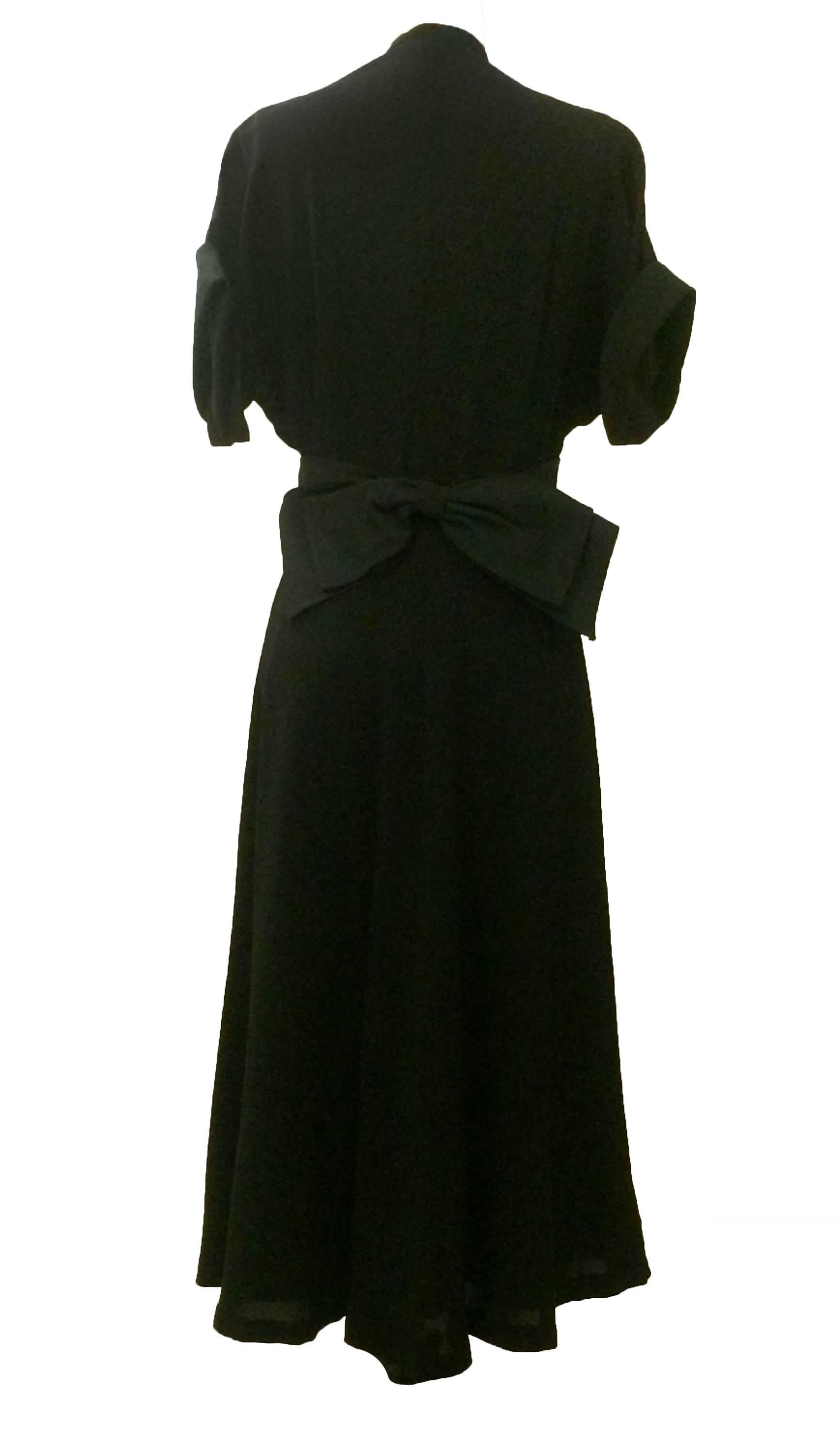 Late 1940's dress from American designer Nettie Rosenstein in black crepe. Pleating at bust, black heavy-faille sash with bow at back, as well as black faille trim at sleeve cuffs. Falls at mid-calf. Side zip. Lined at skirt. 

Lovely drape at