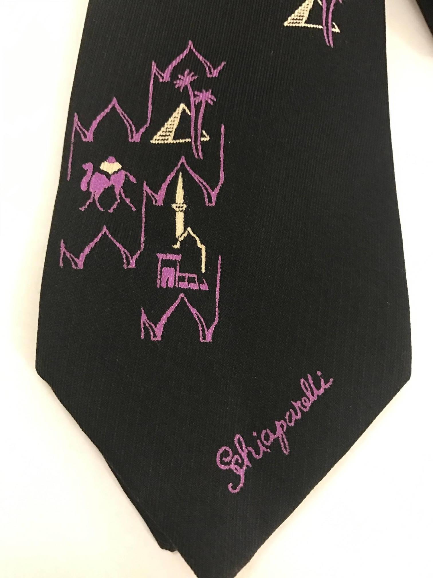 Black neck tie featuring small purple sketches of pyramids, camels, and palm trees. 

Signed 'Schiaparelli' in purple font at bottom front. Unsure of exact decade, but label is from Plum's Inc. boutique in Chicago's legendary Edgewater Beach Hotel,
