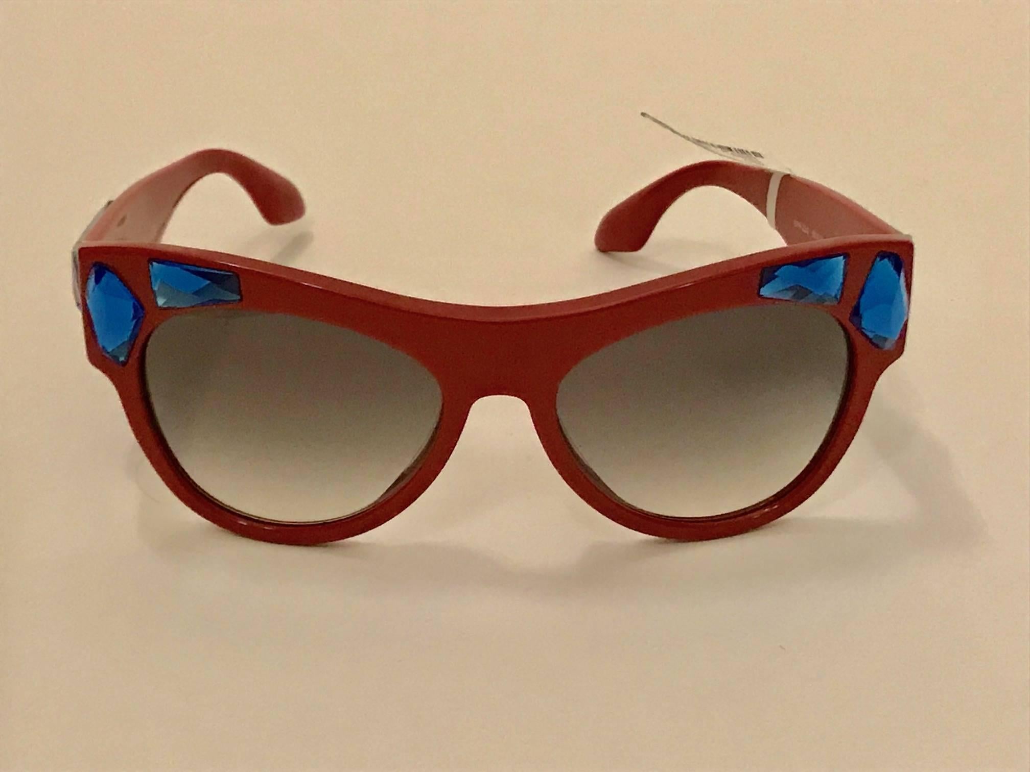 Prada 'Voice' red acetate sunglasses with blue crystal embellishment and gold Prada logo at temple. Grey gradient lenses with 100% UVA & UVB protection.

Size: 140 mm x 56 mm.

Made in Italy.

Excellent condition. New with tags, no flaws to