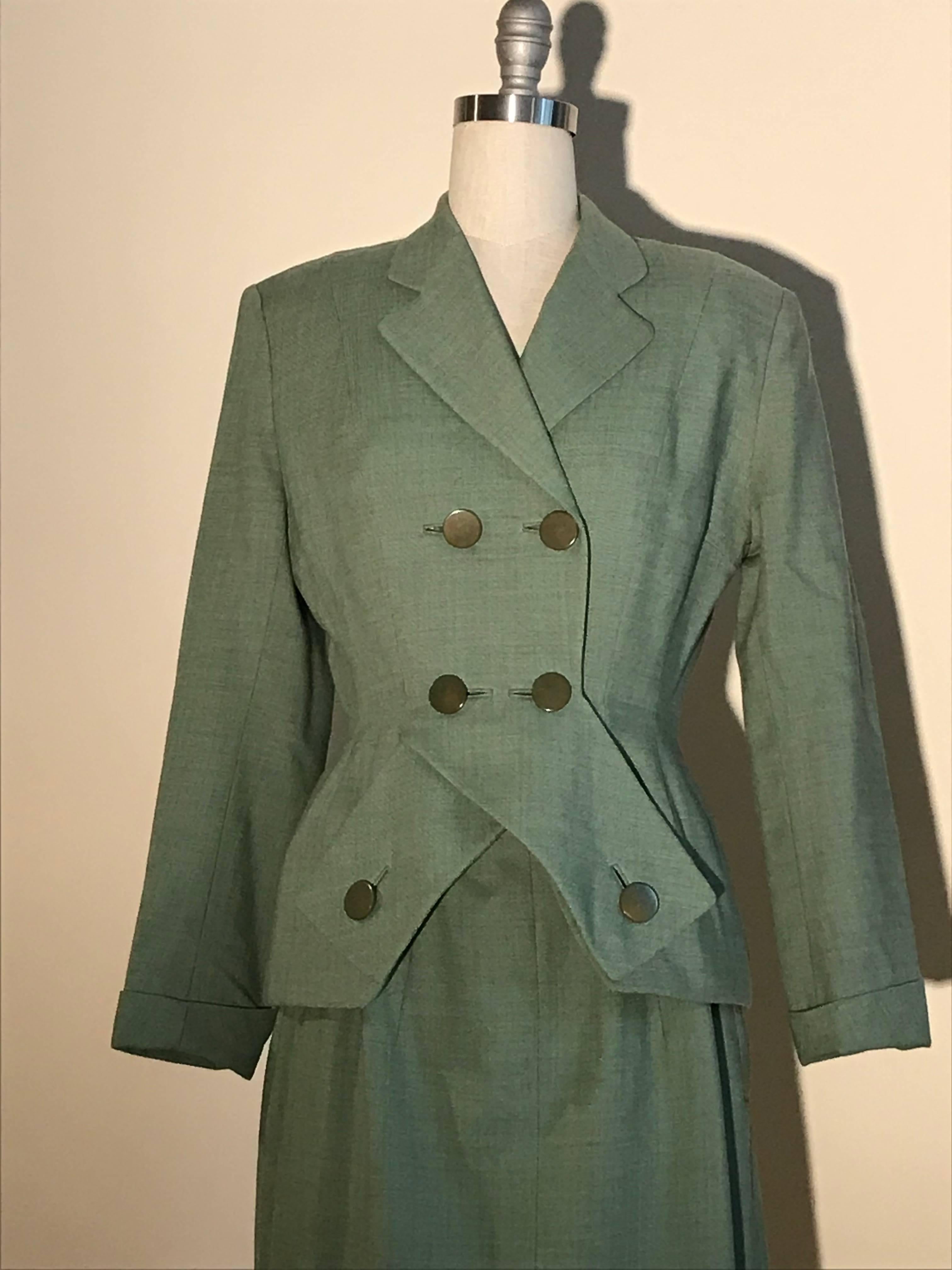 Schiaparelli sage green midi length skirt suit, estimated late 1940s, early 1950s. Criss cross panel at front jacket fastens with mottled green buttons. Light padding at jacket hip creates a New Look shape. Pockets concealed in jacket seam. Cuffed