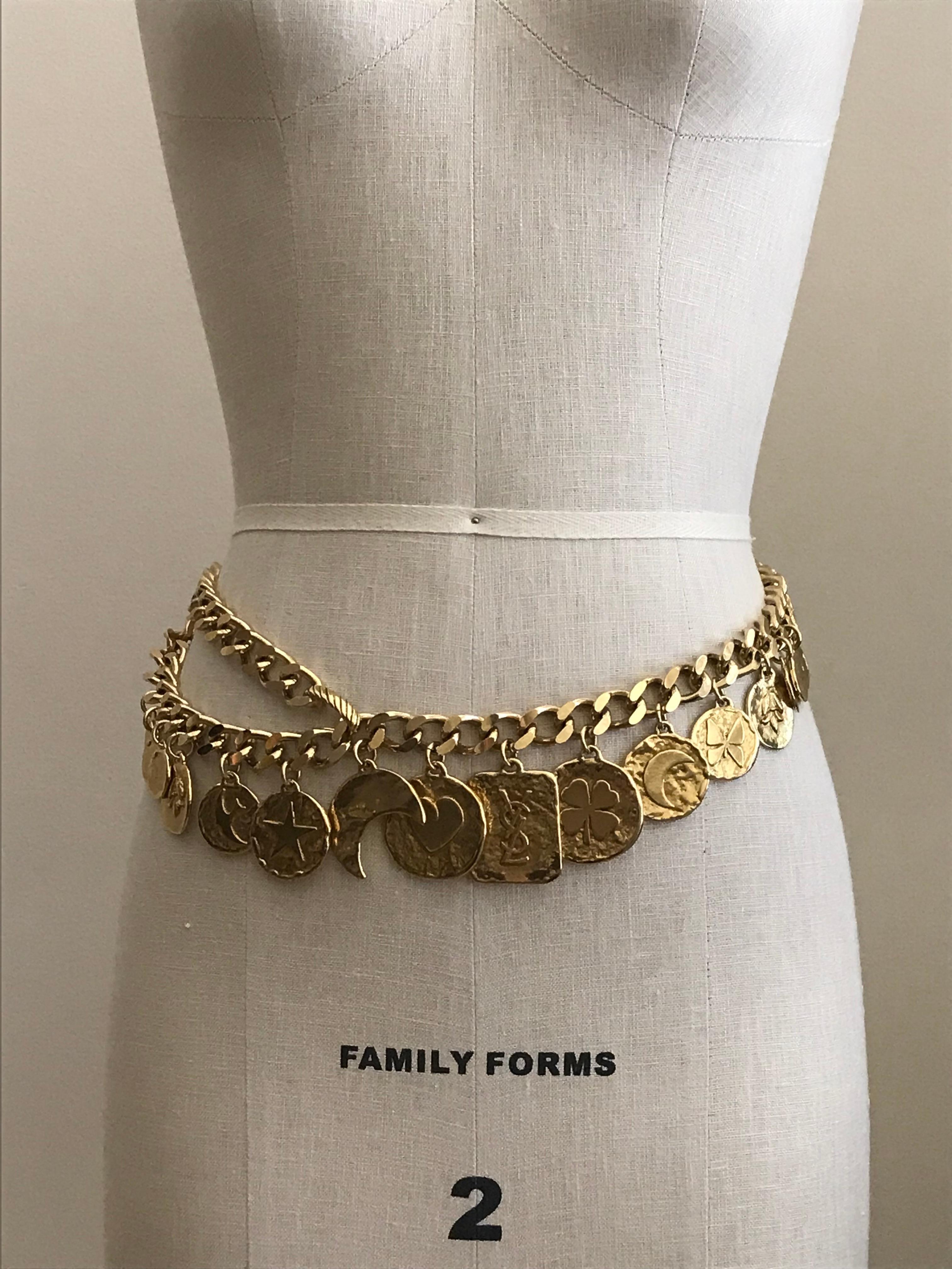 Yves Saint Laurent Collection vintage 1980s gold tone chain link belt with hanging gilt metal good luck charms. Charms include three round blank charms at each side, a fleur-de-lys-like charm at each side, then a dove, star, cut out moon, heart,