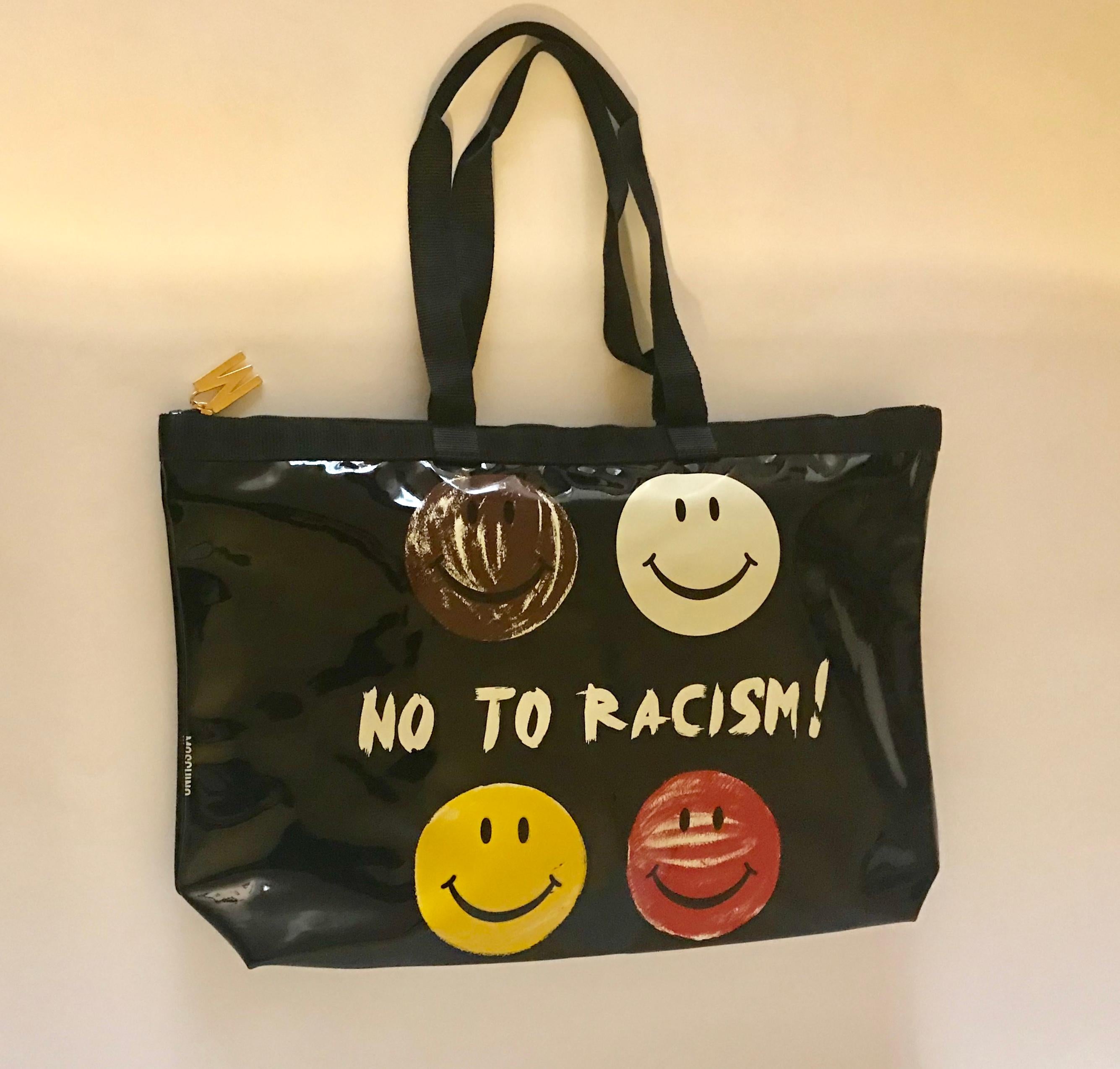 Vintage Moschino by Redwall tote bag in a black patent leather like material (possibly synthetic) featuring multi-colored smiley faces and the slogan 'No To Racism.' Grosgrain trim and handles. M logo at zip top. Signed Moschino at side and Moschino
