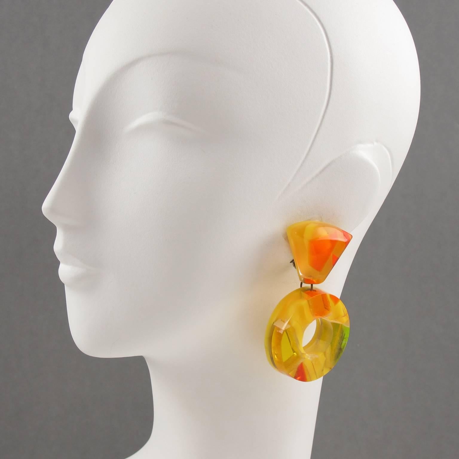 Warm sunny colors oversized lucite clip on earrings designed by Harriet Bauknight for Kaso. Large loop dangling shape with geometric design featuring dimensional multilayers lucite with colorful harlequin inclusions. Contrasted pattern in mirror,