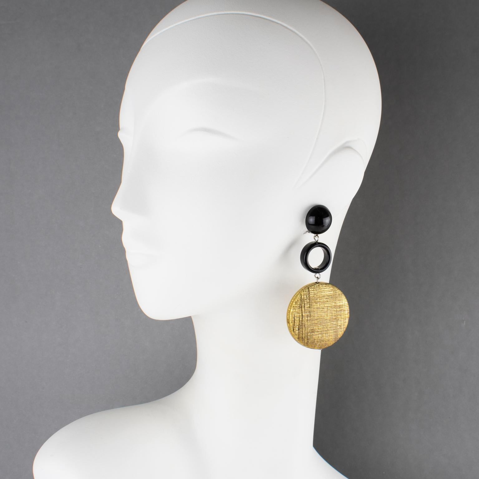 Questi splendidi orecchini a clip in resina e metallo di Angela Caputi, made in Italy, presentano una forma penzolante con elementi geometrici in resina nera contrastati da un importante disco rivestito di metallo color oro con un motivo