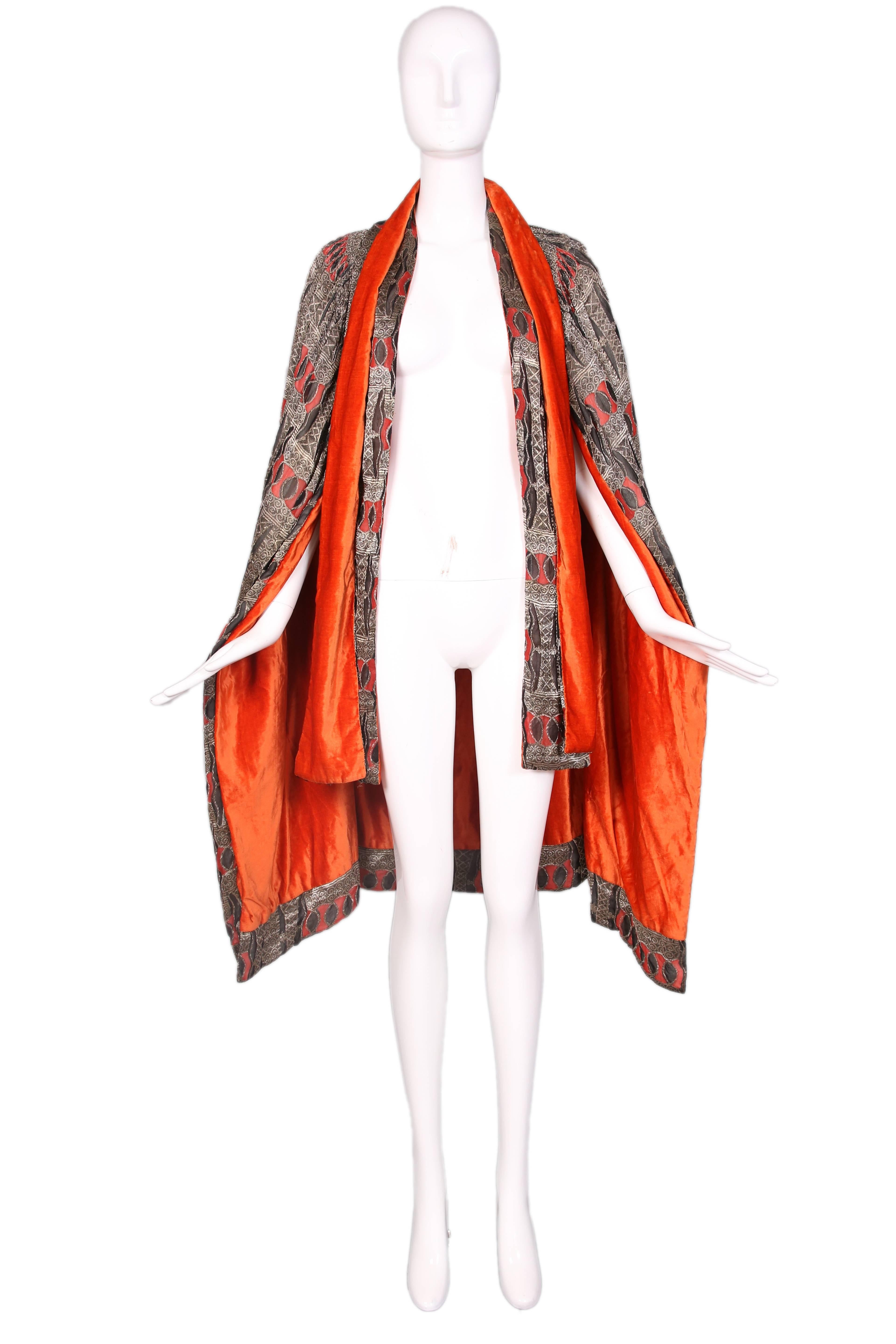 1920's geometric print cape made from gold metallic thread, black and red silk and an interior of orange silk velvet. Cape features two sets of neck ties that drape down the front. Cape isn't labeled but style, design and print reminiscent of Paul