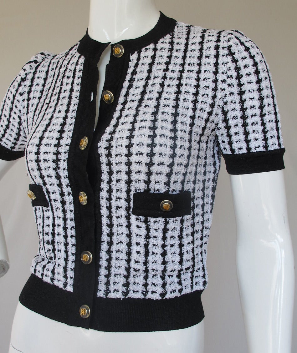 A Chanel black and white boucle short-sleeved cardigan with clear Chanel logo buttons that fasten at center front and at the front pockets. There is no fabric tag but due to its stretch and metallic black trim, my guess is it is a lurex blend. The