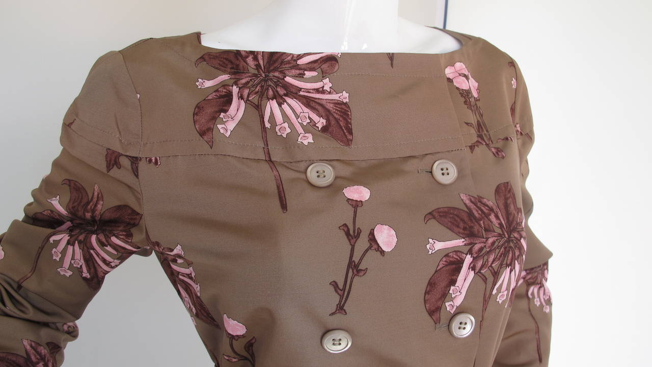 A Prada 100% silk double-breasted light weight coat with a brown/taupe background and floral print in pink and mauve. Features button and snap closure, bateau neckline, dual side pockets, inverted pleat at back and detachable belt. Lined in silk. In