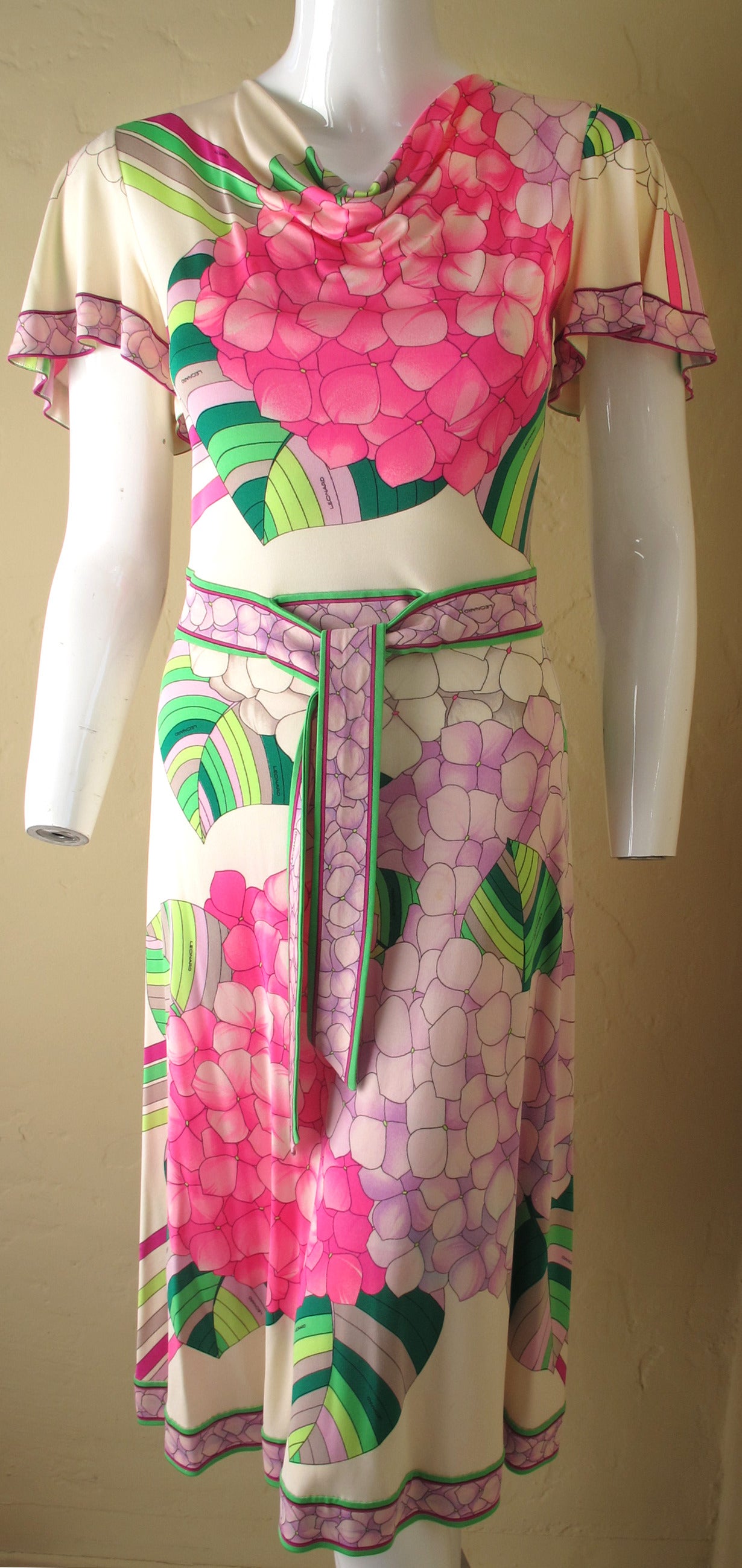 Unusual knee-length day dress by Parisian designer, Leonard made from silk jersey. Dress features a cowl neckline, flutter sleeves, two frontal ties at the waste and a graphic floral print in various shades of pink, green, lilac, fuchsia, gray and