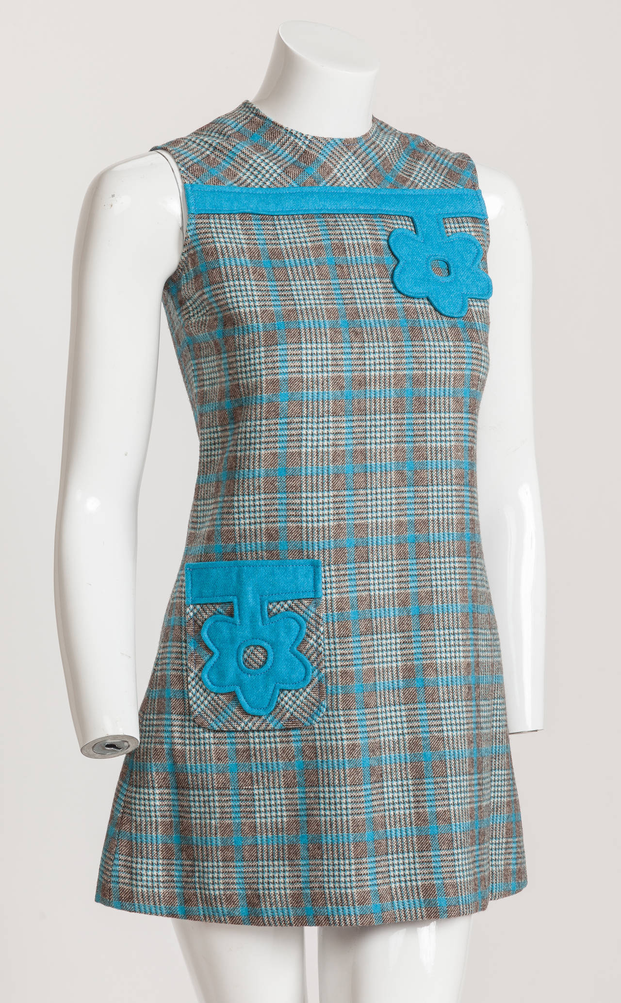 A Pierre Cardin sleeveless blue, brown and creme plaid wool mini dress lined with light purple Pierre Cardin signature fabric. Most likely from the 1970s, this tiny mod mini dress is appliquéd with two oversize teal blue felt flower and bar designs