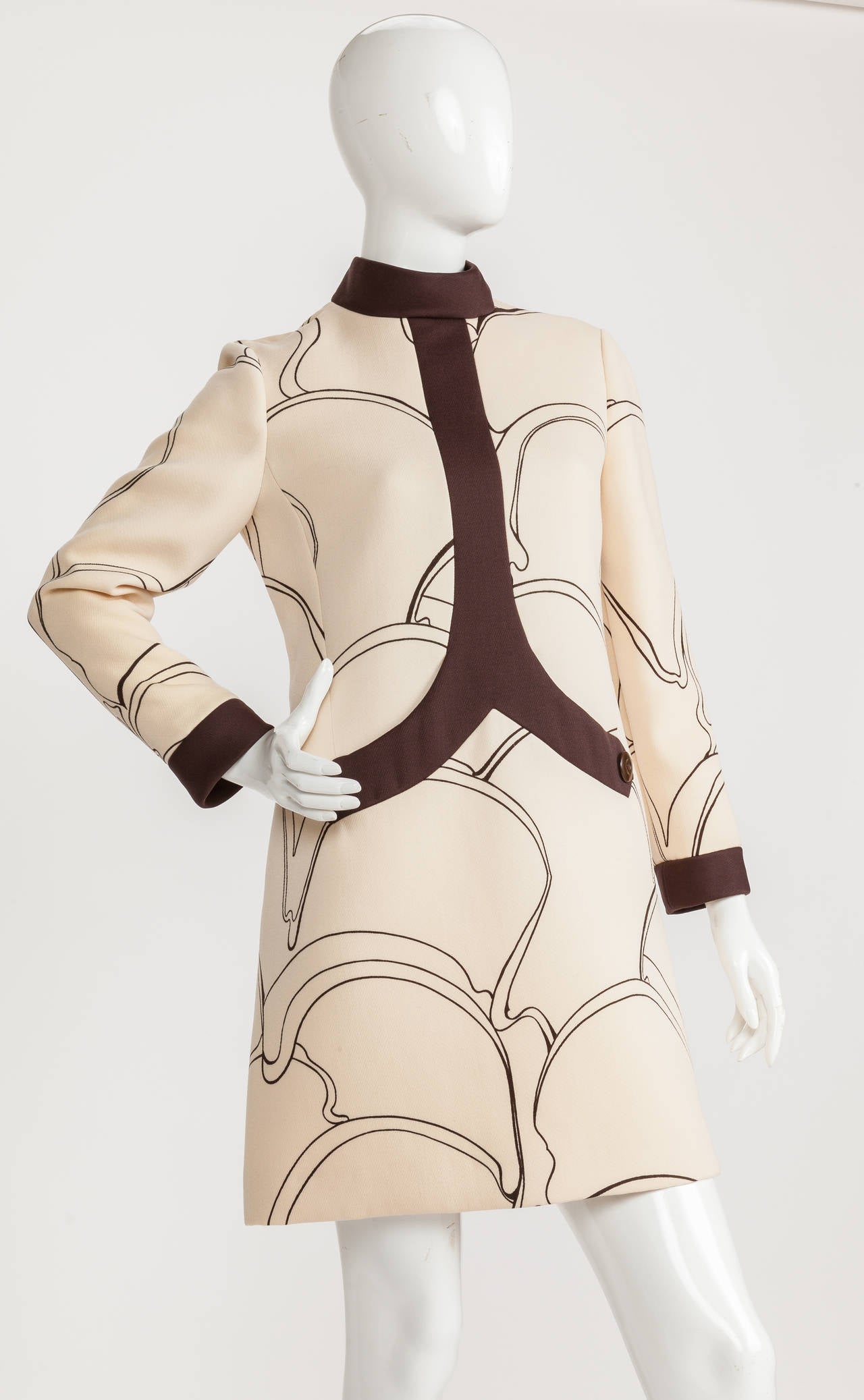 A 1969 Pierre Cardin wool mini-dress with an abstract organic print in brown on a creme background and a contrasting brown band at the standing collar that bisects the bodice and finishes in two curved pocket bands and button trim. Also features