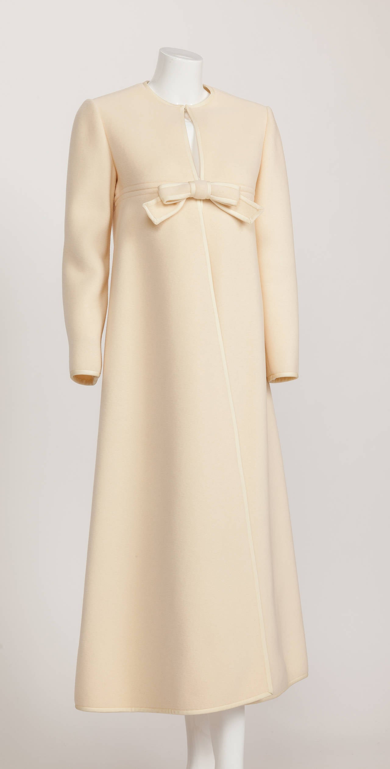 A 1960's Pierre Cardin creme-colored melton wool overcoat with bow and silky trim at the edges. The A-line silhouette is created by means of a diagonally cut front flap that fastens via hook and eye closure at the neck and behind bow. There is a