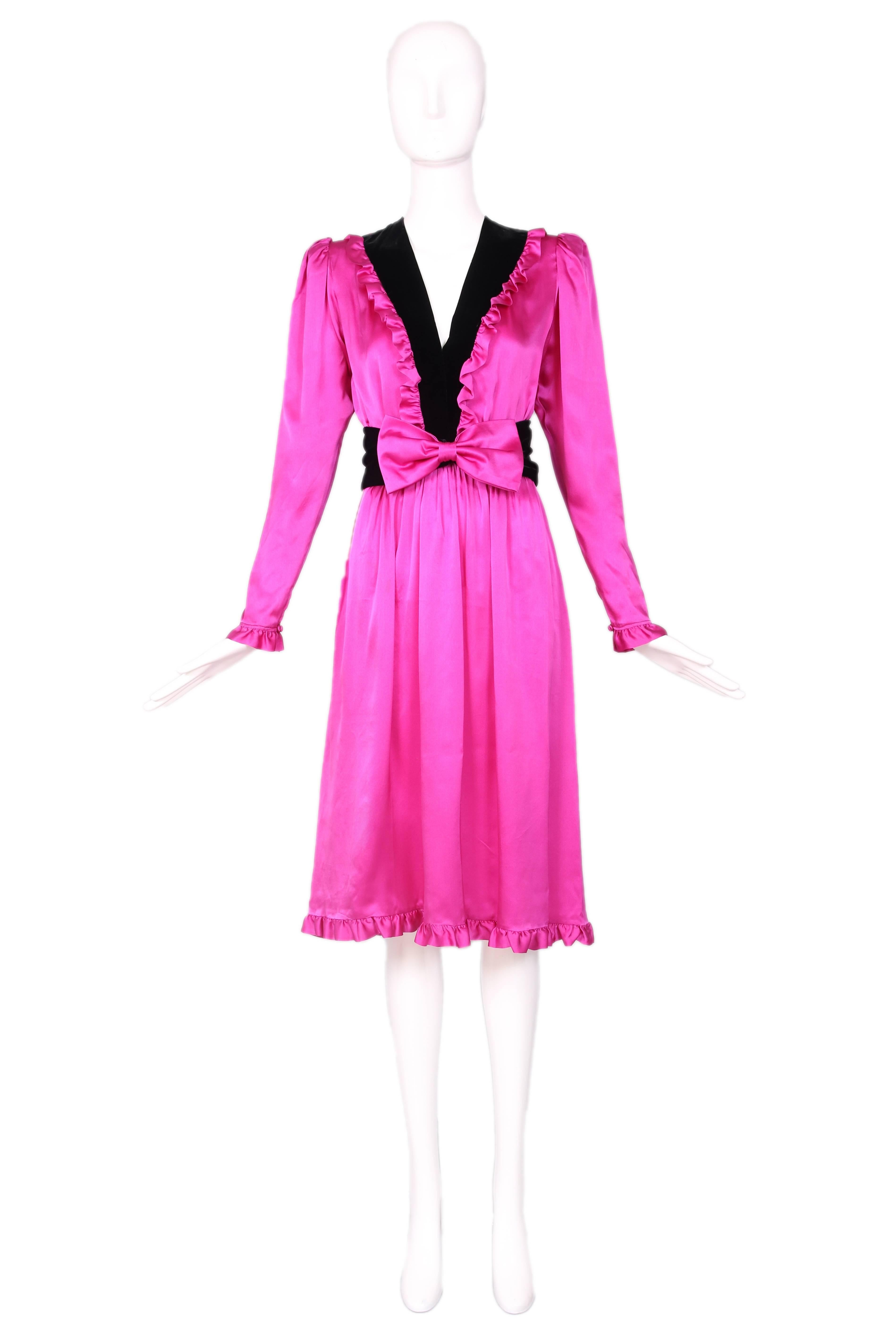 A 1970s Yves Saint Laurent fuschia silk day dress with a black velvet trimmed deep V-neckline as well as ruffled trim at the hem, sleeves and velvet neckline detail. Comes with a detached black velvet belt with oversized fushcia silk bow. Hidden