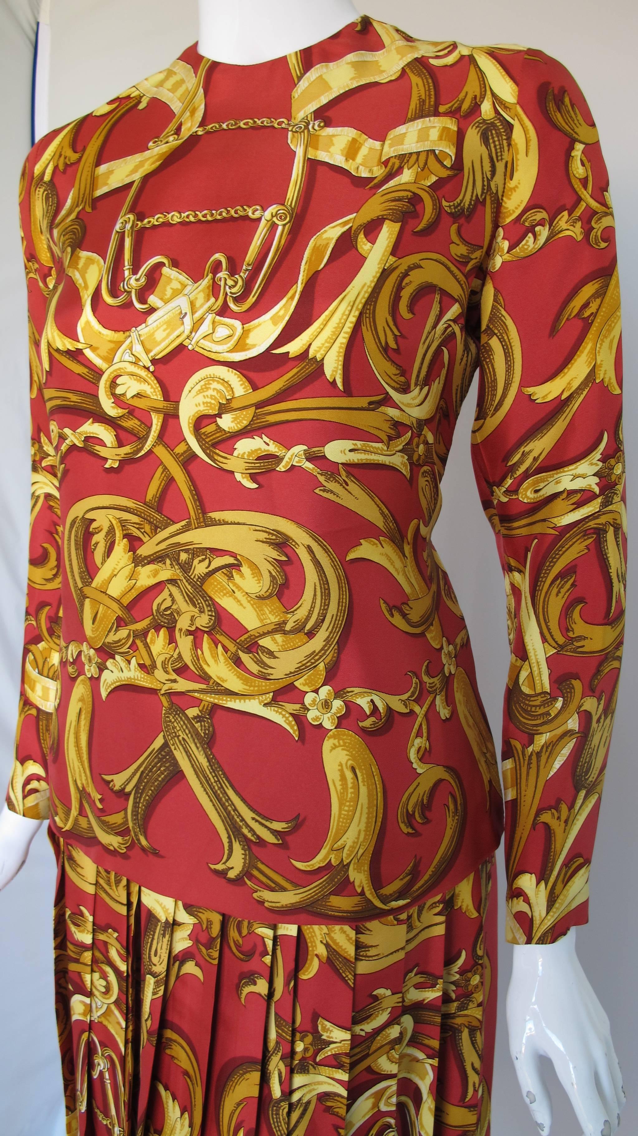 A vintage Hermes 100% silk blouse and skirt ensemble in a classic Hermes print in shades of red, yellow and gold. Ensemble consists of a long sleeved blouse that zippers at the back neck and the cuffs as well as a long pleated skirt with one hidden