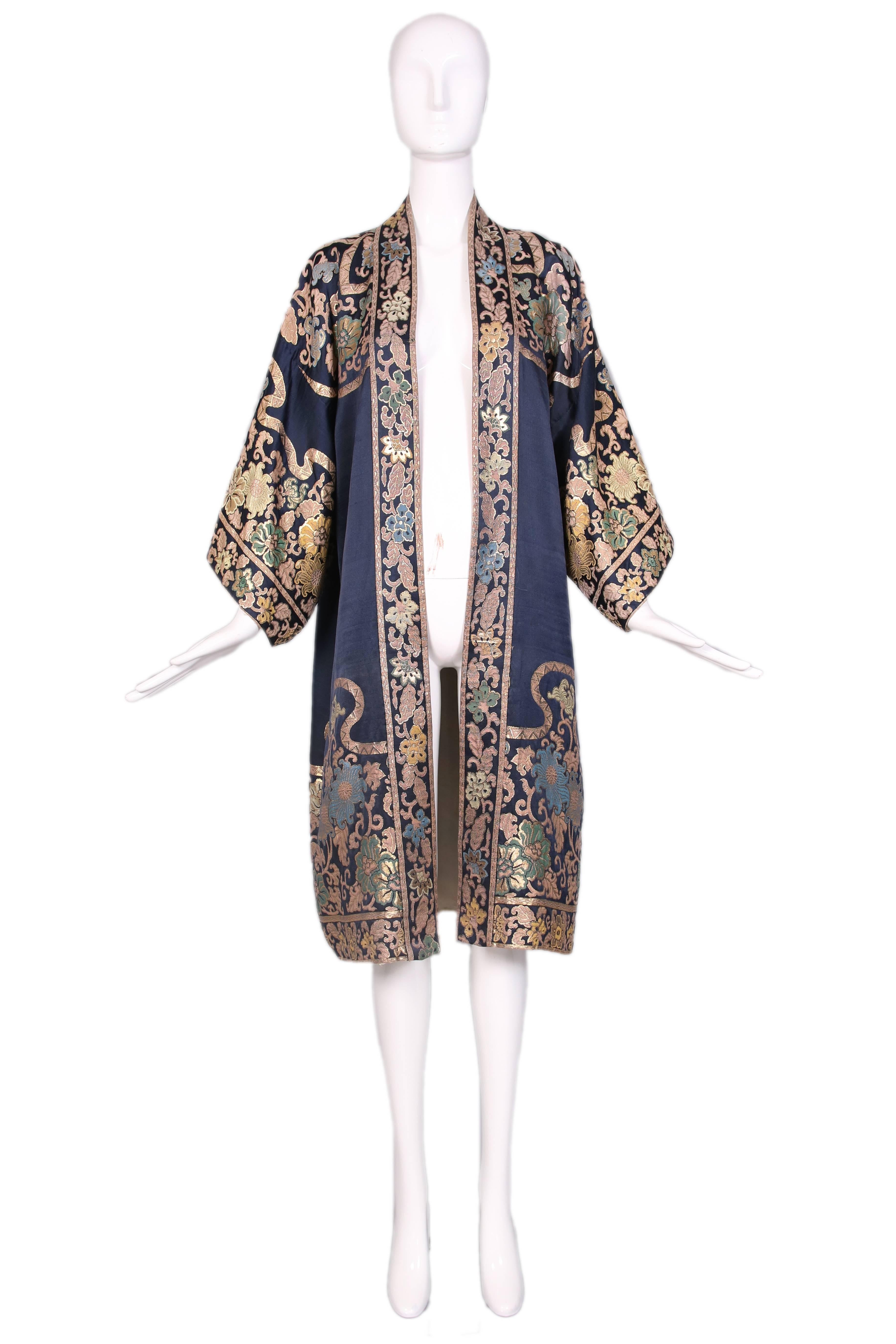 1920's blue silk open front coat duster with metallic floral motif and kimono sleeves. Fully lined in silk. In very good to excellent condition with some slight, scattered yellow stains at the interior lining and two nickel sized areas at the back
