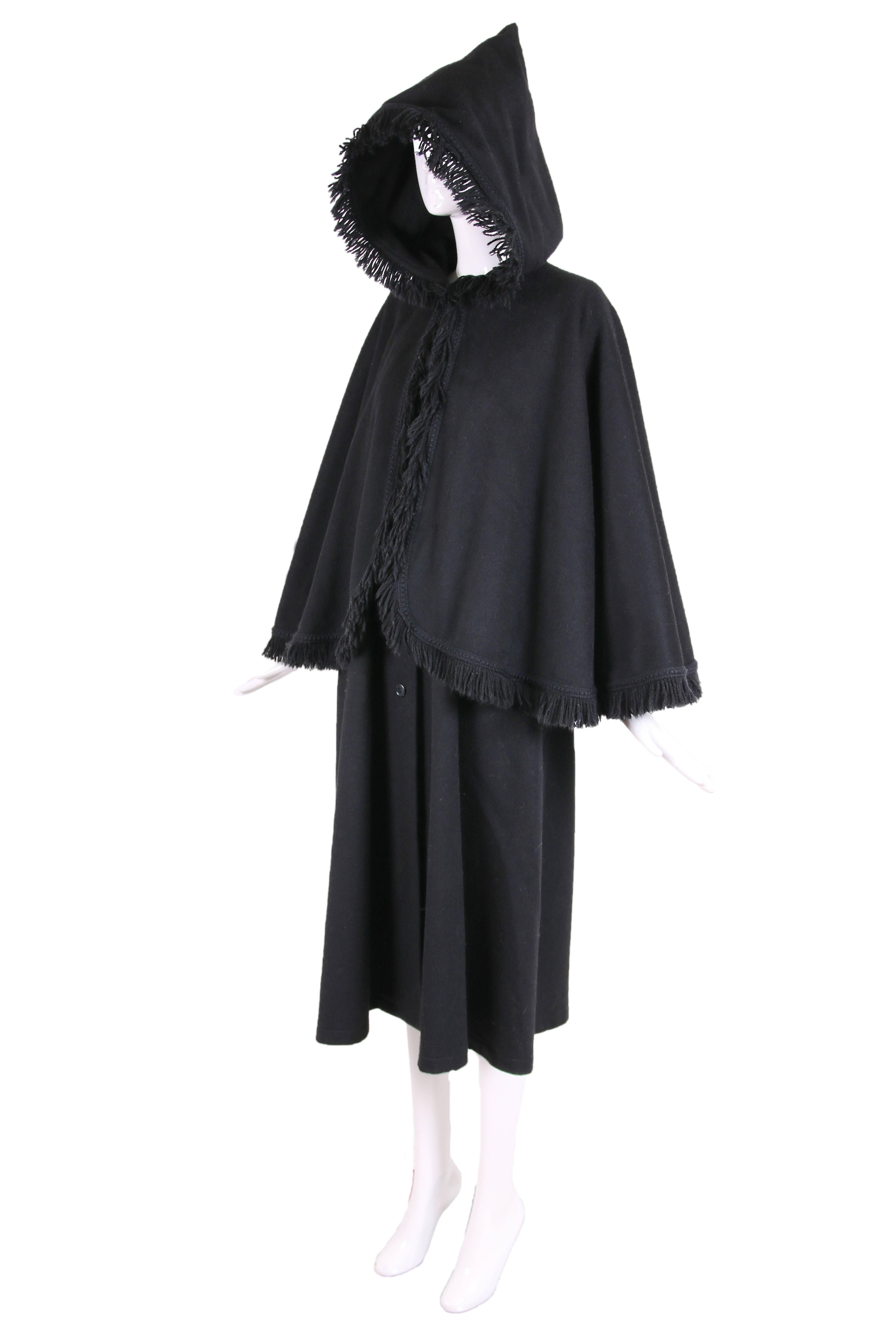 1970's Yves Saint Laurent black wool cape coat with hood and wool fringe trim. Coat underneath cape has a swing silhouette, button closure down center front and hidden side pockets. In excellent condition with a tiny hole in the interior fabric