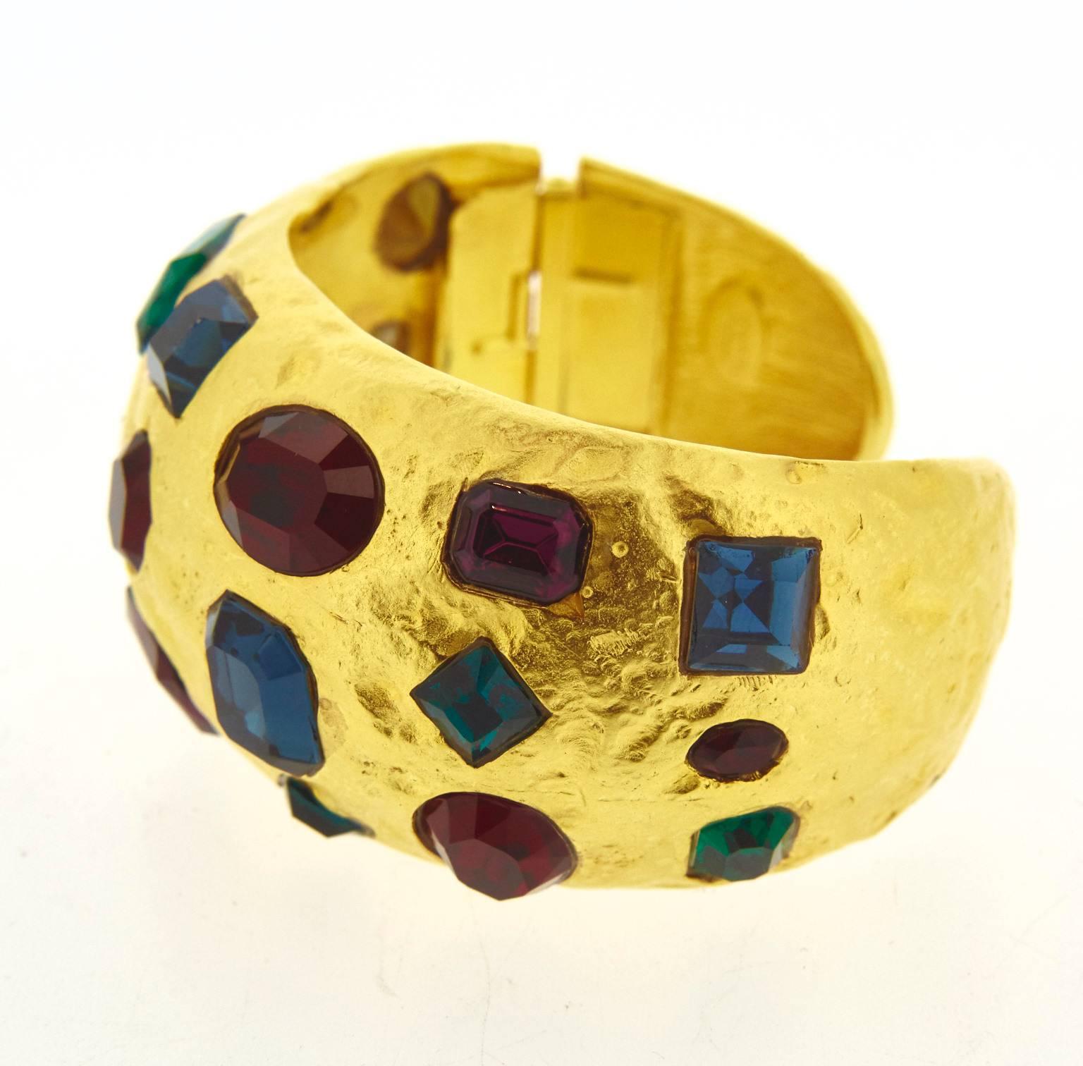 A gold plated clamper bracelet by Kenneth Jay Lane New York. Set with multi crystals in purple, emerald green, sapphire blue and red. One end has a clamper hinge opening up to put the bracelet on.

It measures 3.4cm high by 5.6cm wide, 17.5cm 7