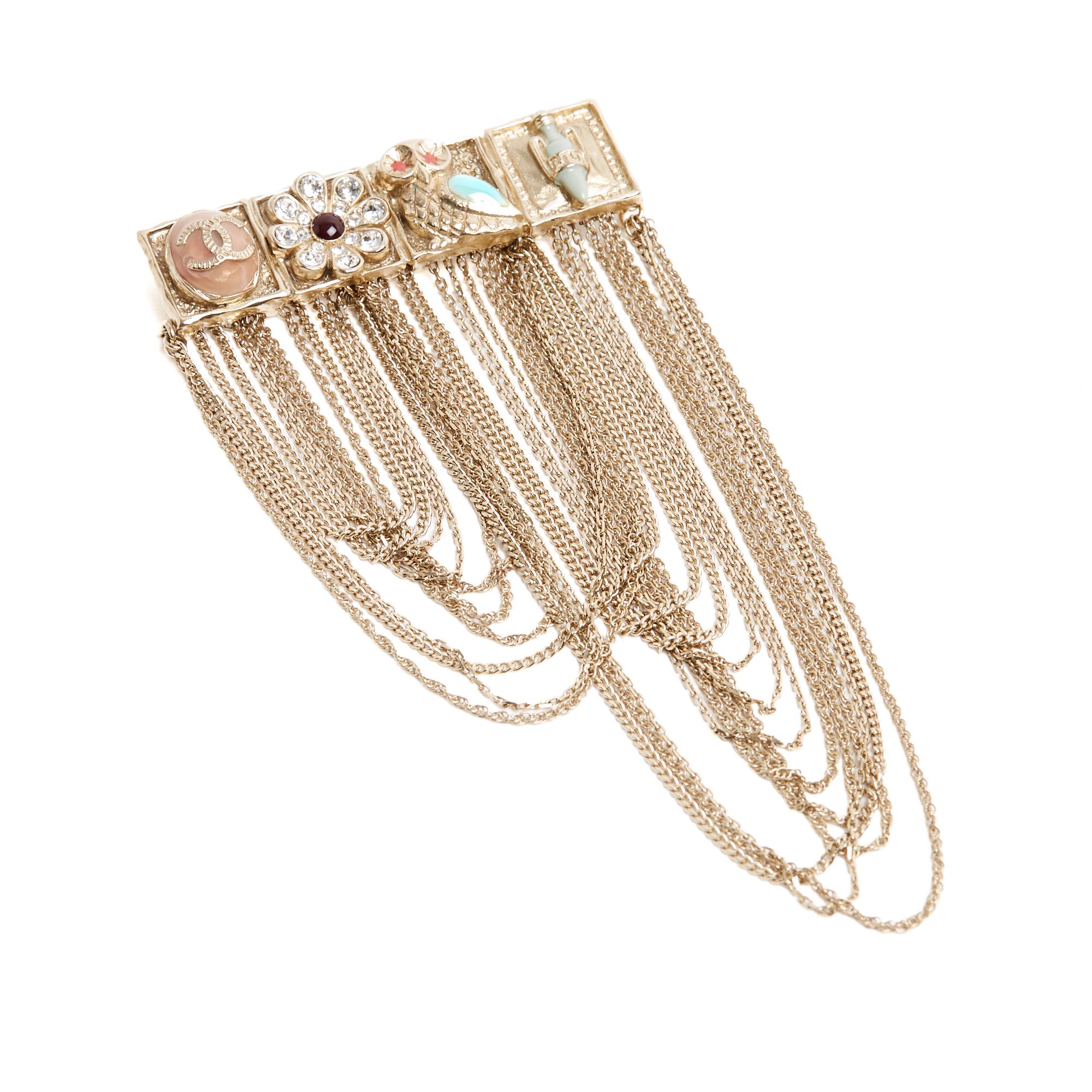 Broche de la collection Chanel Cruise 2018 (sur le thème de la Grèce antique) en métal doré pâle composée d'une rangée de 4 motifs - un logo CC sur un cabochon émaillé rose, une fleur incrustée de strass et d'émail bordeaux, un hibou aux yeux en