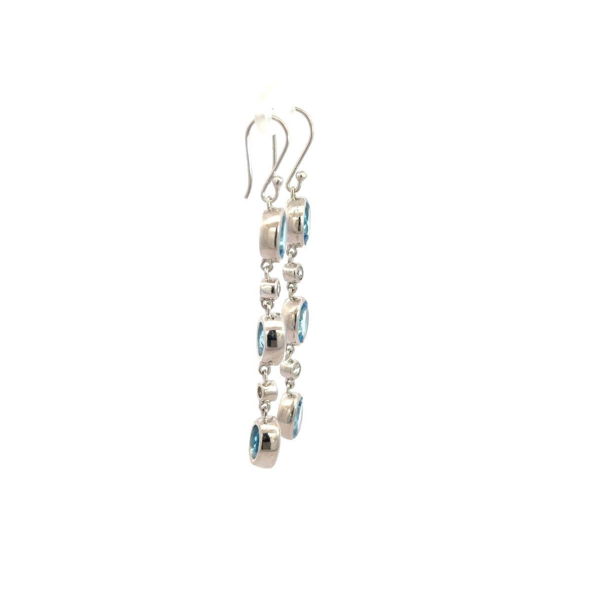 Ces boucles d'oreilles pendantes linéaires avec topaze bleue et zircon sont fabriquées en argent 925 lumineux et présentent un trio captivant de pierres précieuses topaze bleue taillées en ovale, chacune sertie en biseau pour une silhouette nette et