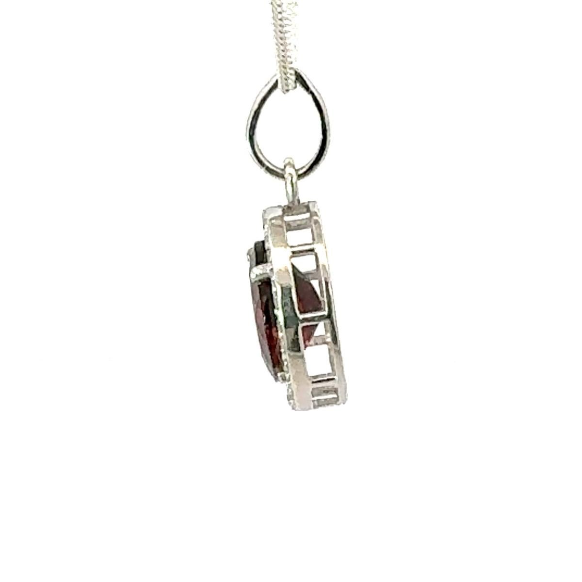 Ce pendentif en argent 925 est orné d'un grenat ovale d'un rouge profond. Entouré d'un délicat halo de zircon cubique scintillant, le pendentif est suspendu à une attache et à une chaîne en argent, rayonnant à la fois de raffinement et d'éclat.