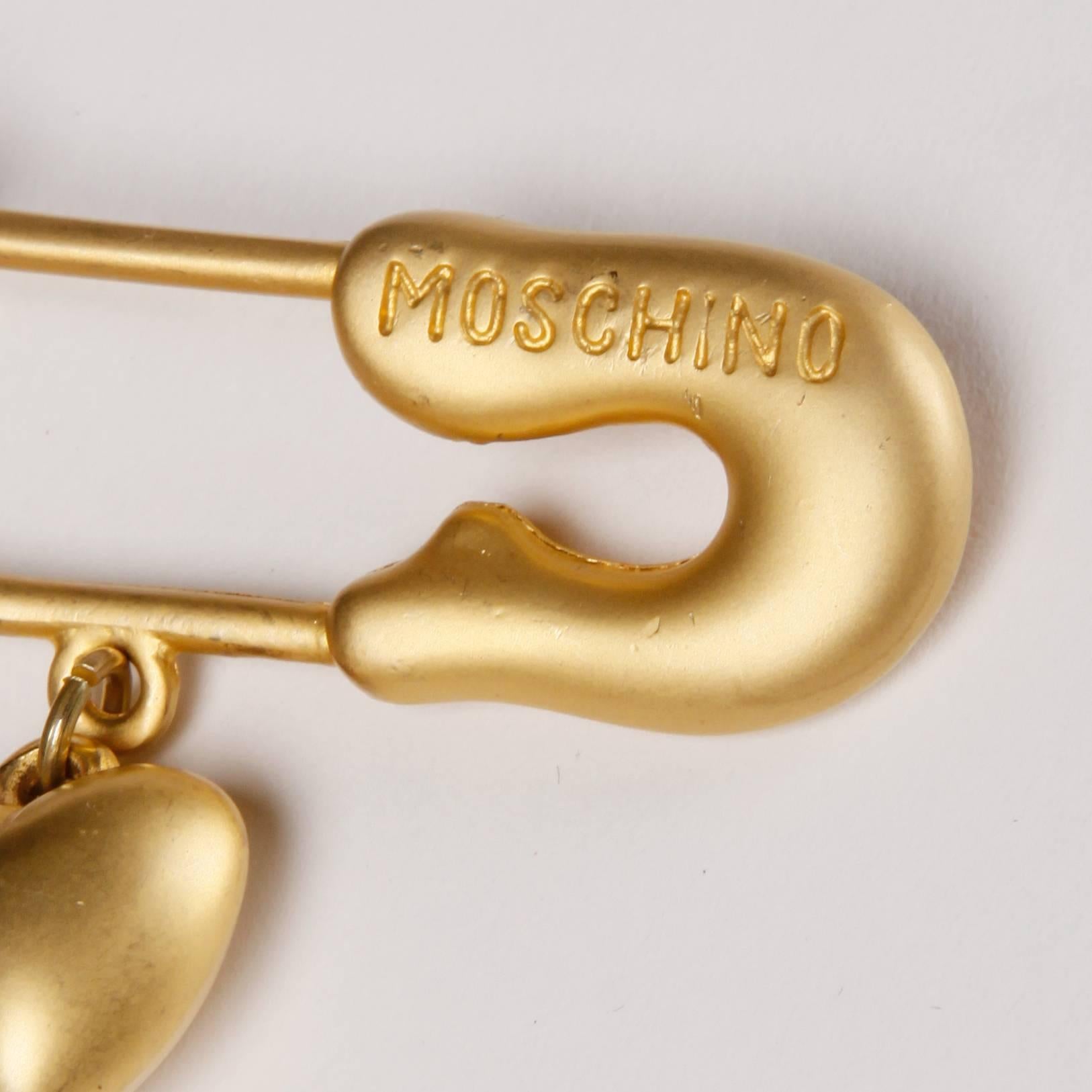 Vintage Moschino gold brooch in the shape of an oversized safety pin. The brooch has three dangling charms; a peace sign, an ampersand, and a heart. From Moschino's iconic 
