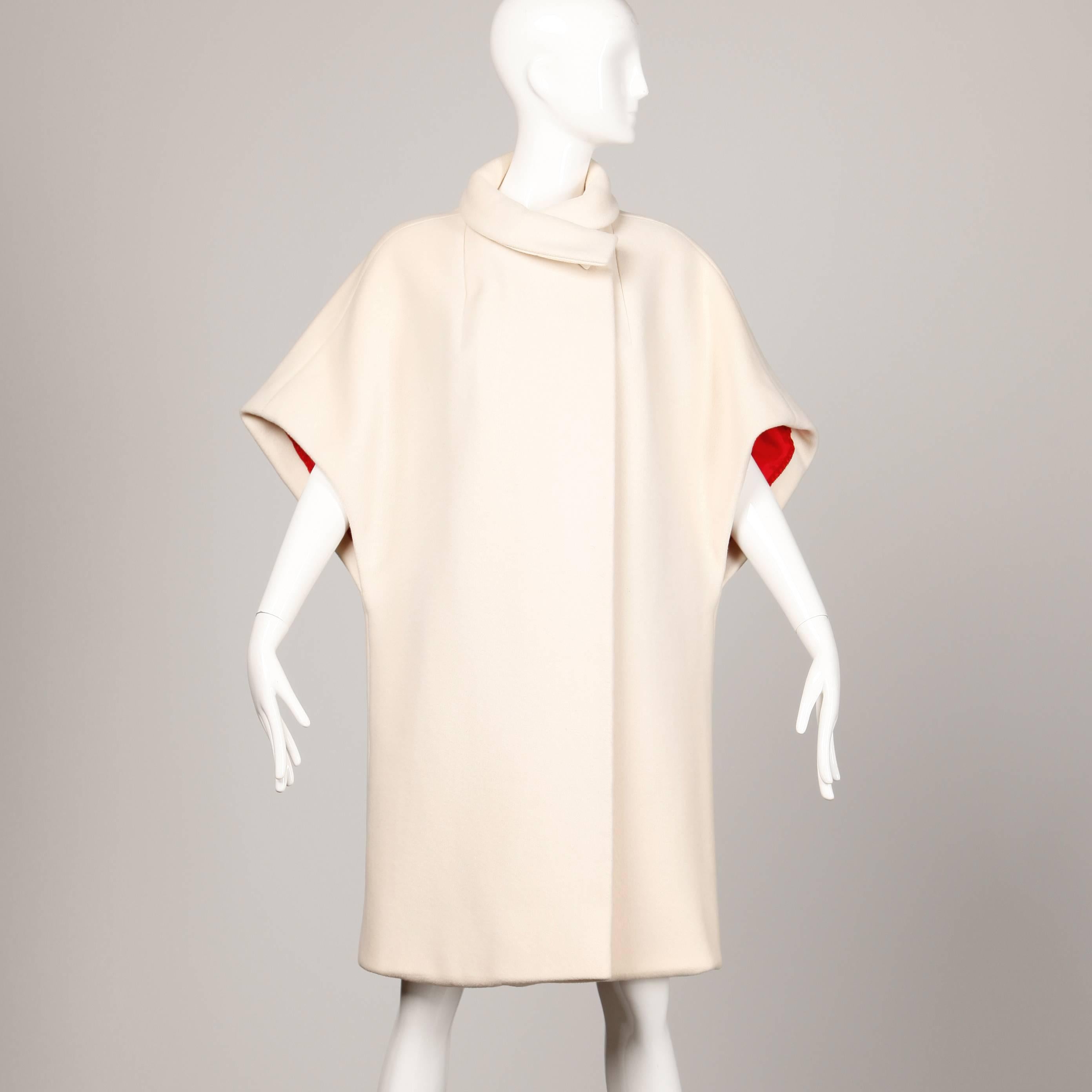 Stunning 1960s cape coat in ivory wool by Via Veneto for Milgrim. Cherry red lining and asymmetric front button closure. The marked size is 10 and the coat fits like a modern size small-medium (forgiving fit). The measurements are as follows: Bust-