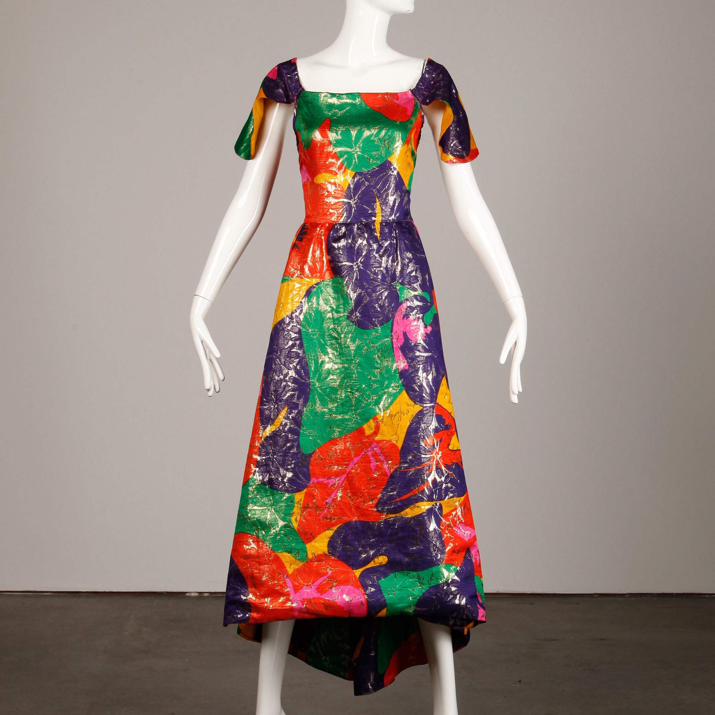 Incroyable robe vintage en tissu métallisé d'Arnold Scaasi avec un ourlet haut et bas et un dos ouvert avec des chaînes. Manches à rabat en forme de pétales et tissu de soie coloré imprimé floral d'inspiration tropicale. Entièrement doublé, il se