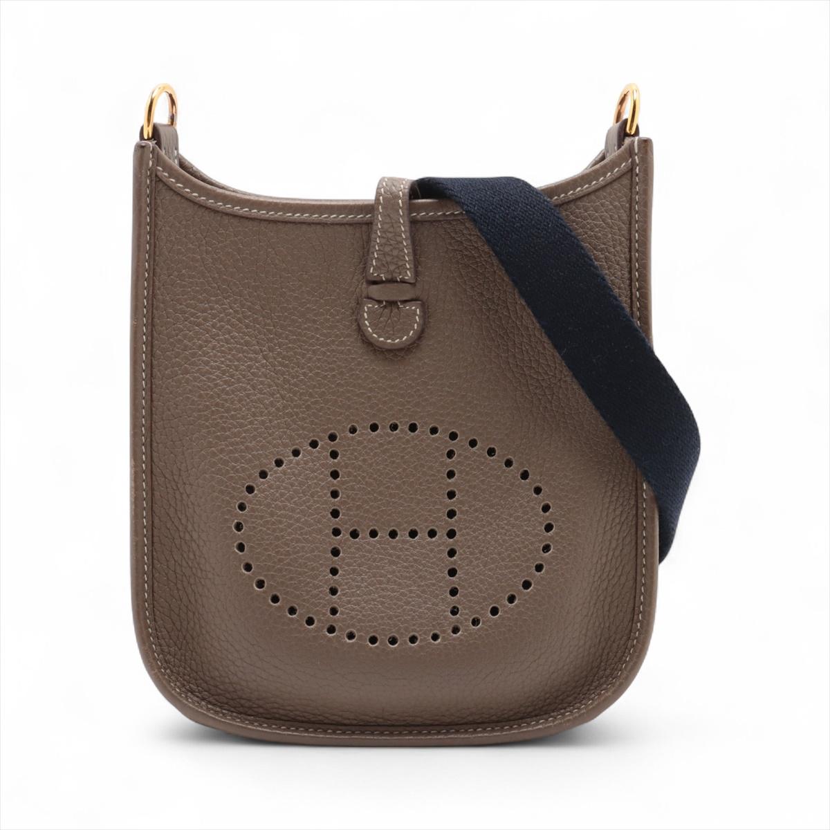 El bolso Hermès Evelyne 16 TPM Amazone de piel Etoupe Taurillon Clémence es un bolso bandolera compacto y práctico que ejemplifica el lujo discreto. Confeccionado en piel Clémence flexible y de grano grueso en el versátil tono Etoupe, incorpora el