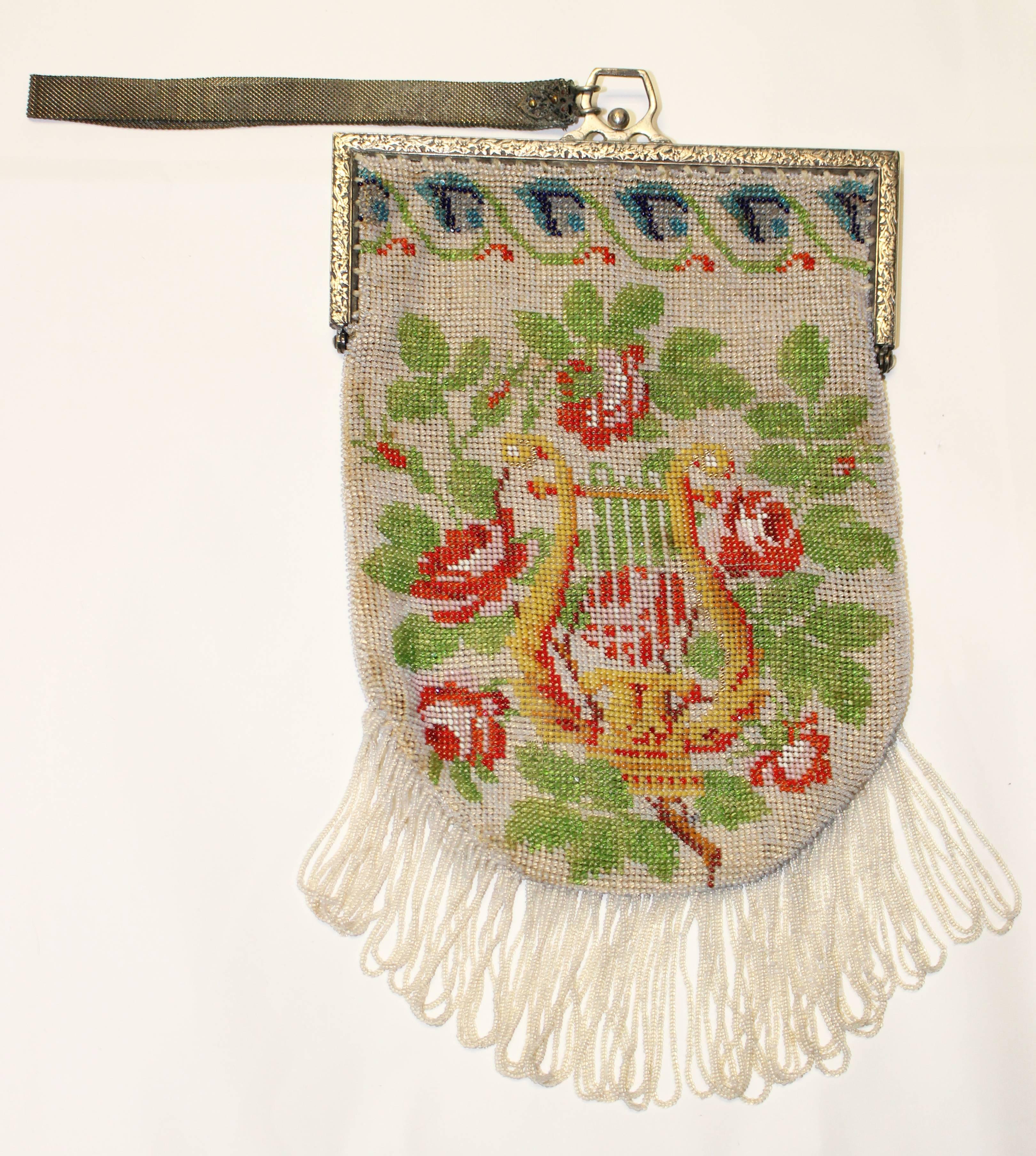 handgekettelte Handtasche aus den 1920er Jahren mit grünen Blättern, roten Rosen und einer Harfe auf einem weißen Feld aus Saatperlen. Blaue Blumen umrahmen den oberen Rand. Silberfarbener geprägter Rahmen. Unbeschriftet. Geschlungene