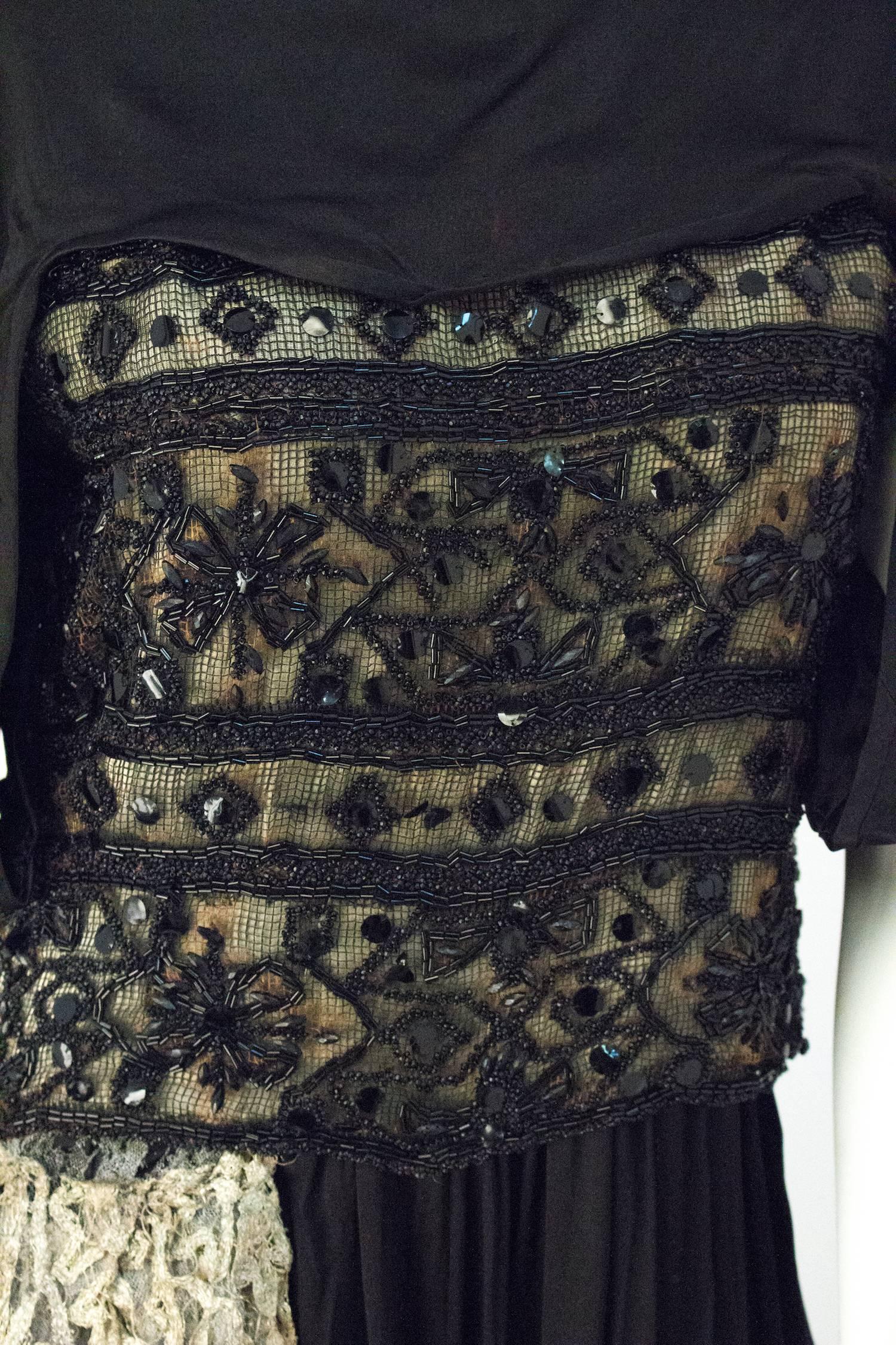 Robe de soirée édouardienne en soie noire avec dentelle lamée et perlage argentés. La jupe se noue à la cheville sur le devant gauche de la jupe. Embellissement en dentelle lamée argentée le long de la cuisse droite, avec des ornements en pompons