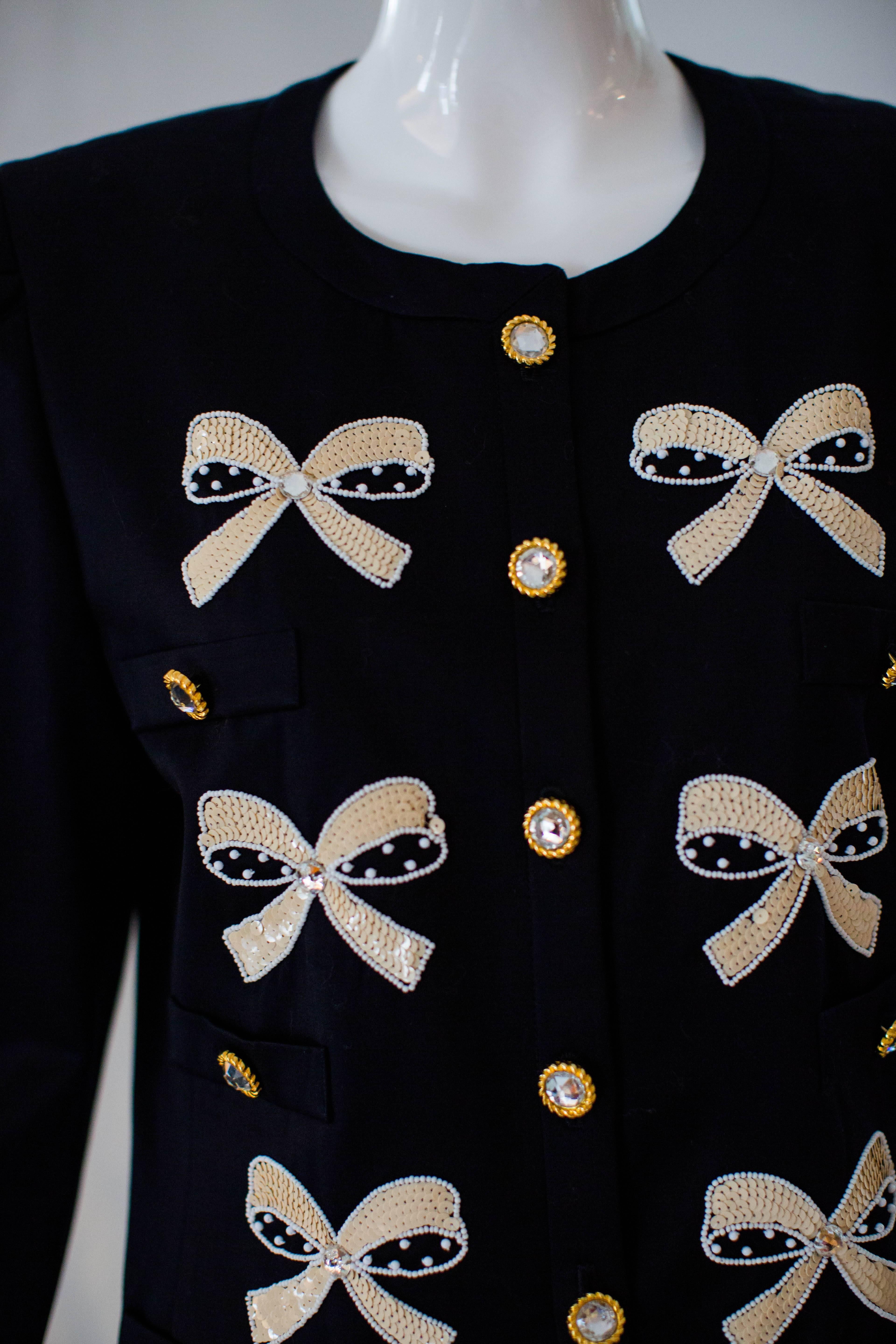 1990s vintage Escada jacket in navy, with a collarless design, long sleeves, darted detail at the shoulder cap, and sequin beaded bows that adorn the front of the jacket with contrasting rhinestone and gold tone buttons. This charming  jacket is a