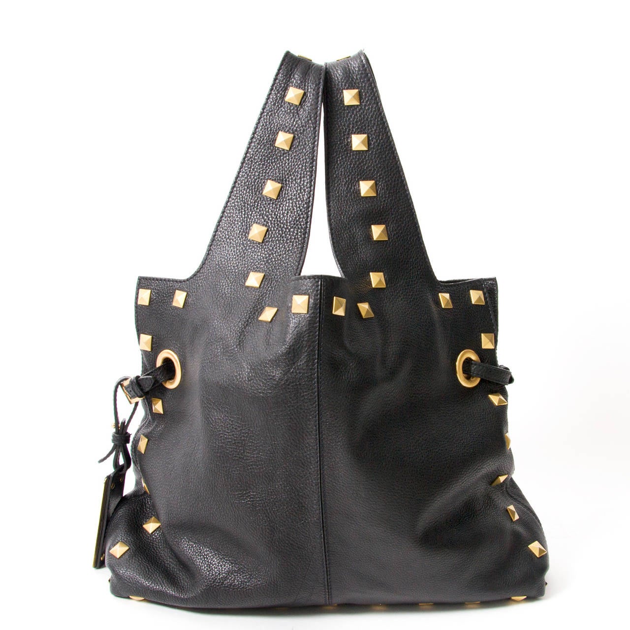 Black shopper with two large top handles in supple leather.

The bag features large gold-tone studs on the handles, sides and on the front in a heart shape.

On each side are two holes with adjustable strap. On one side there is a gold plate