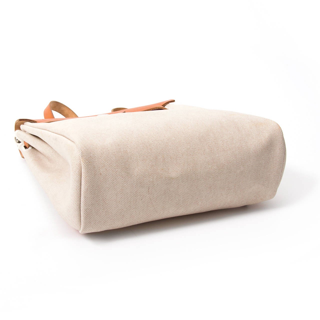 This Hermès Herbag comes in woven white and beige canvas with cognac leather detailing.
It has a noticeable trapezoid shape and a front flap similar to the Kelly bag, but with a different closure. Thanks to the leather straps, you can wear this