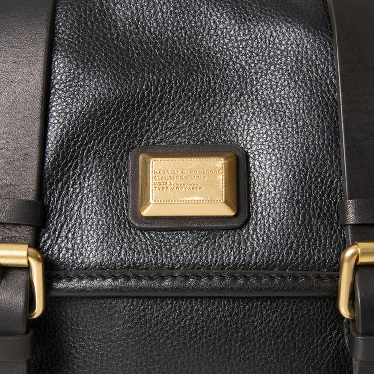 Marc by Marc Jacobs black leather handbag with gold tone hardware. Two straps at the flap with buckle detailing. Two push buttons as closure at the front.

Open pocket beneath the flap.

The interior features a zipped pocket, a cellphone holder