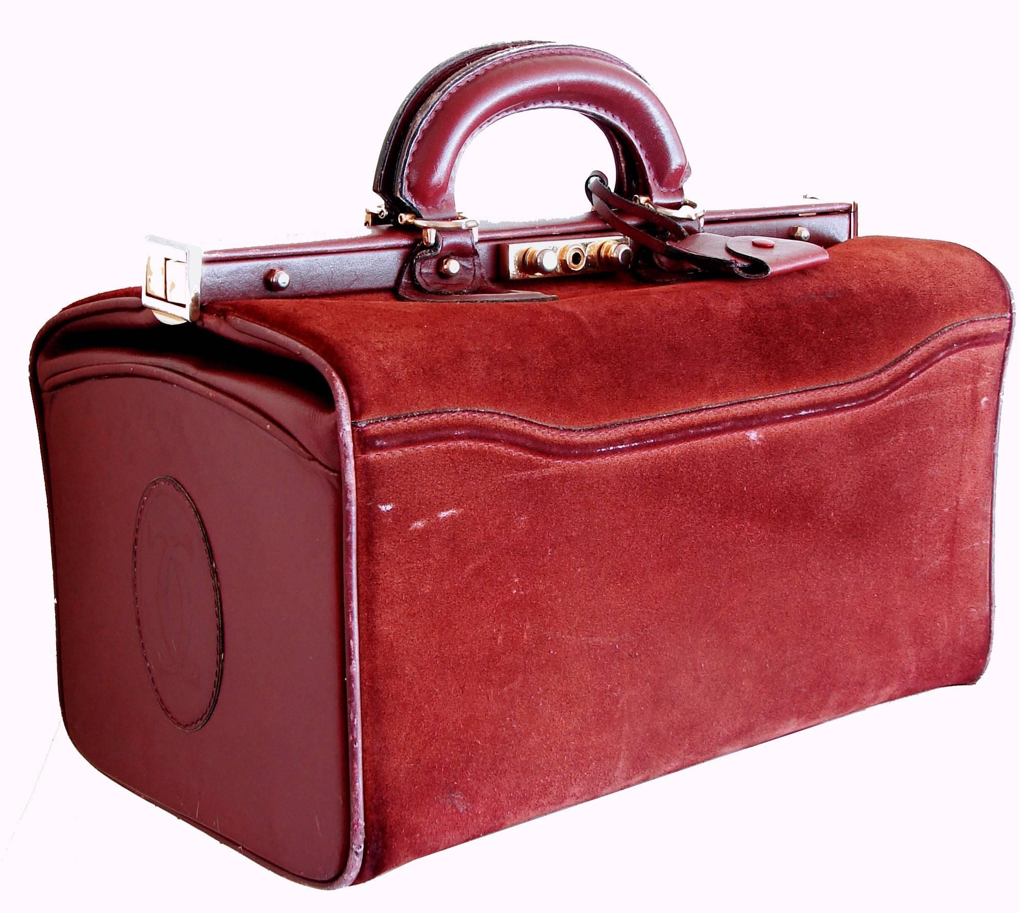 This large travel bag was made by Cartier in the late 1980s.  Made from Bordeaux suede and leather, it features the Cartier logo on each side, a top handle, a push lock fastener with key lock and a matching leather clochette with key.  Inside, its