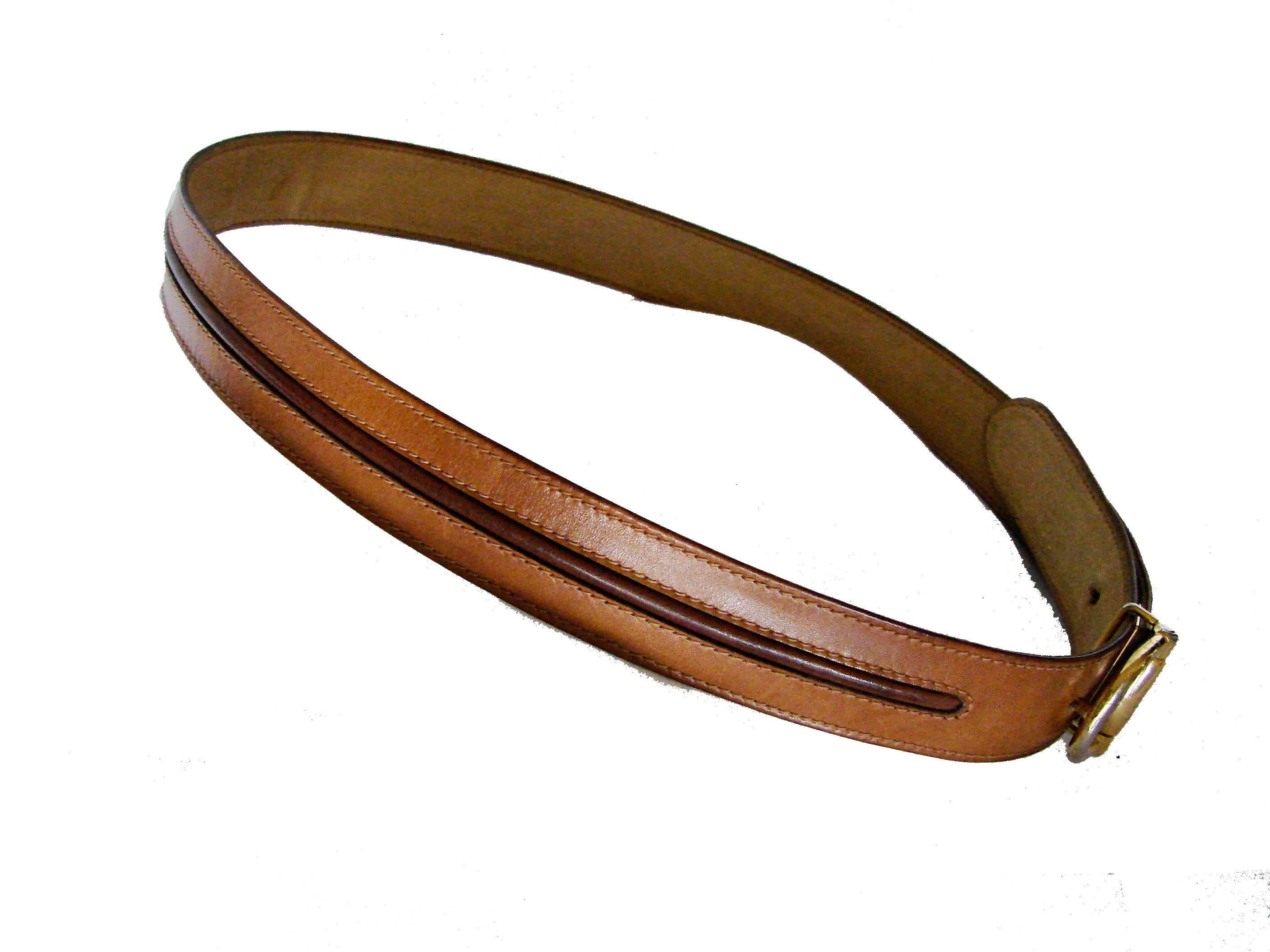 This horse bit buckle and belt was made by Gucci in the late 1970s.  Made from tan leather, the belt strap features a dark brown leather stripe at its center and the horse bit buckle is made from gold tone metal.  In good condition overall, the gold