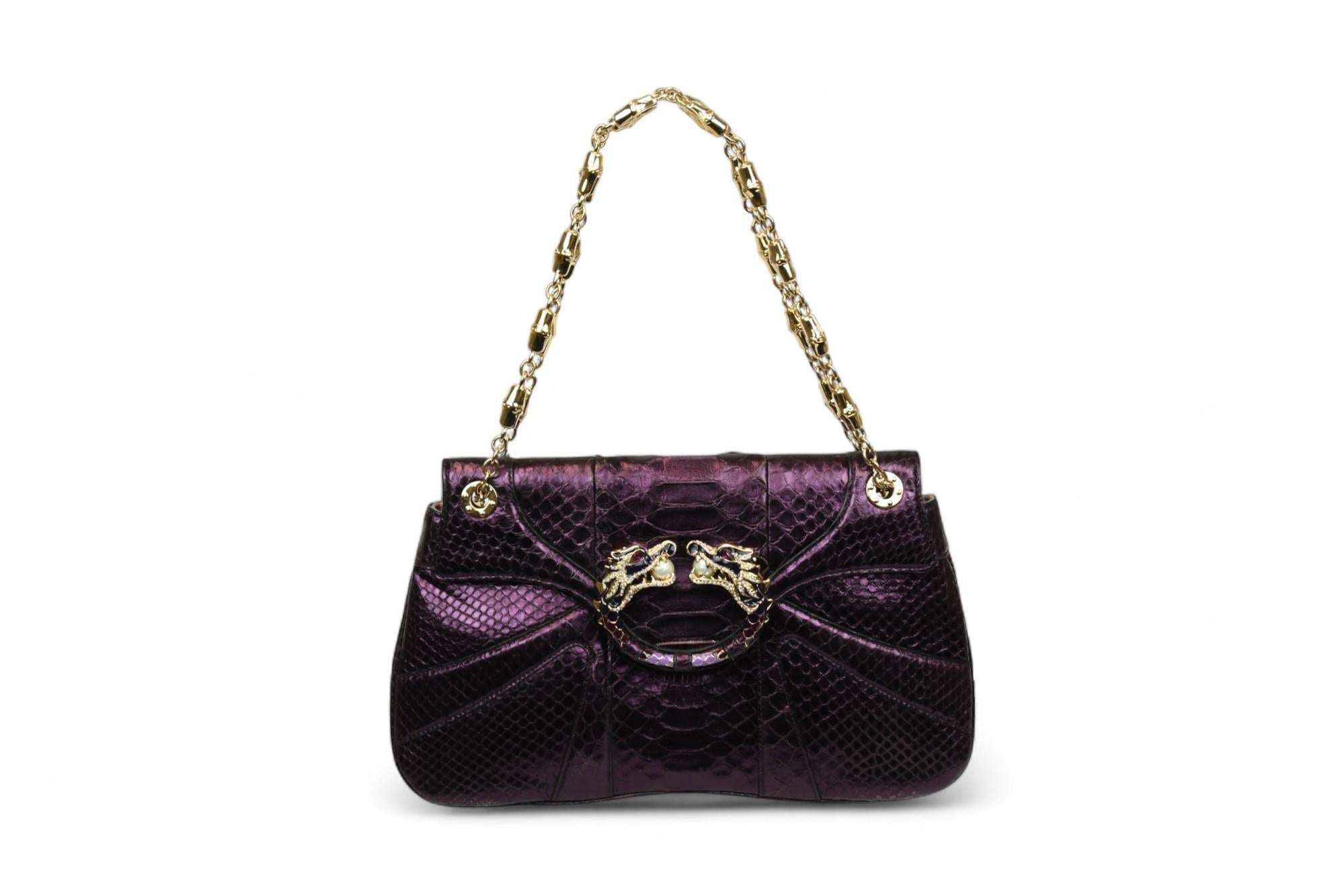 Cette magnifique pochette Roberto Cavalli présente le design audacieux caractéristique de la marque, réalisée dans un luxueux cuir python gaufré violet métallisé. La pièce maîtresse - un double motif de tête de dragon avec des ferrures dorées et des