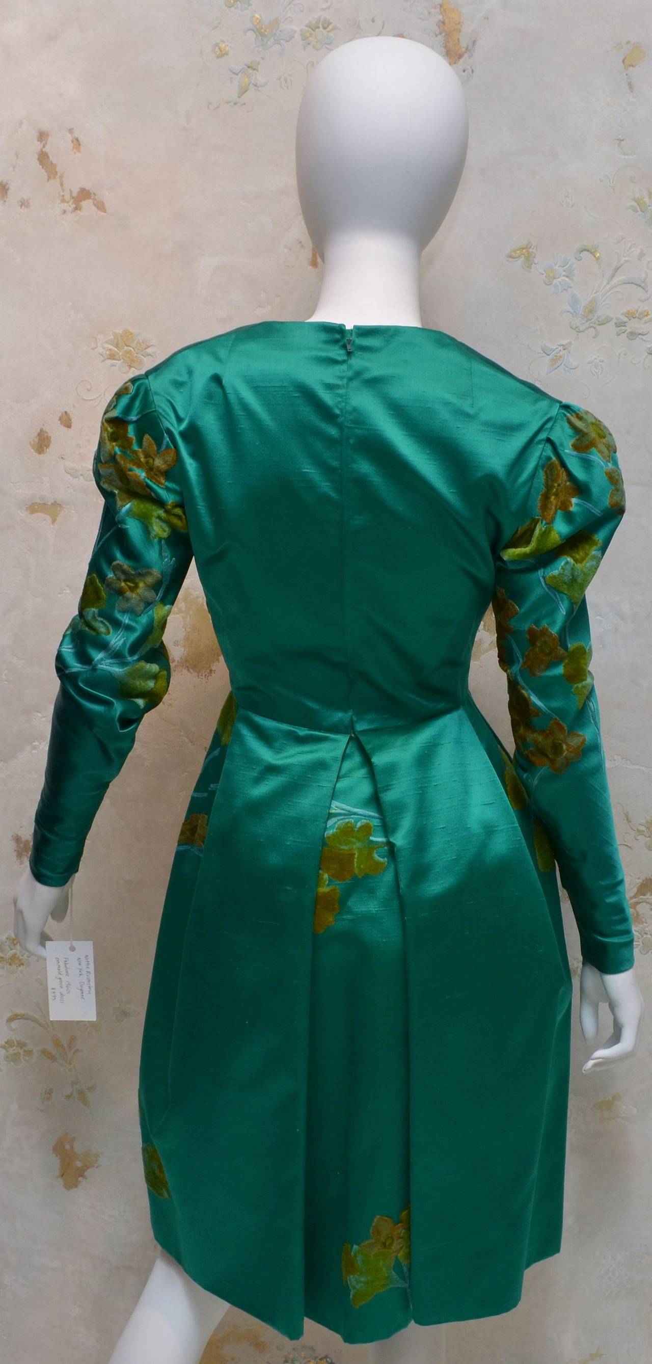 Vintage Nettie Rosenstein dress in emerald green silk satin features a back zipper closure, silk velvet floral detailing, fully lined, and with zippers at the cuffs as well. Note waist seam does not break pattern and flower is cut out and hand sewn