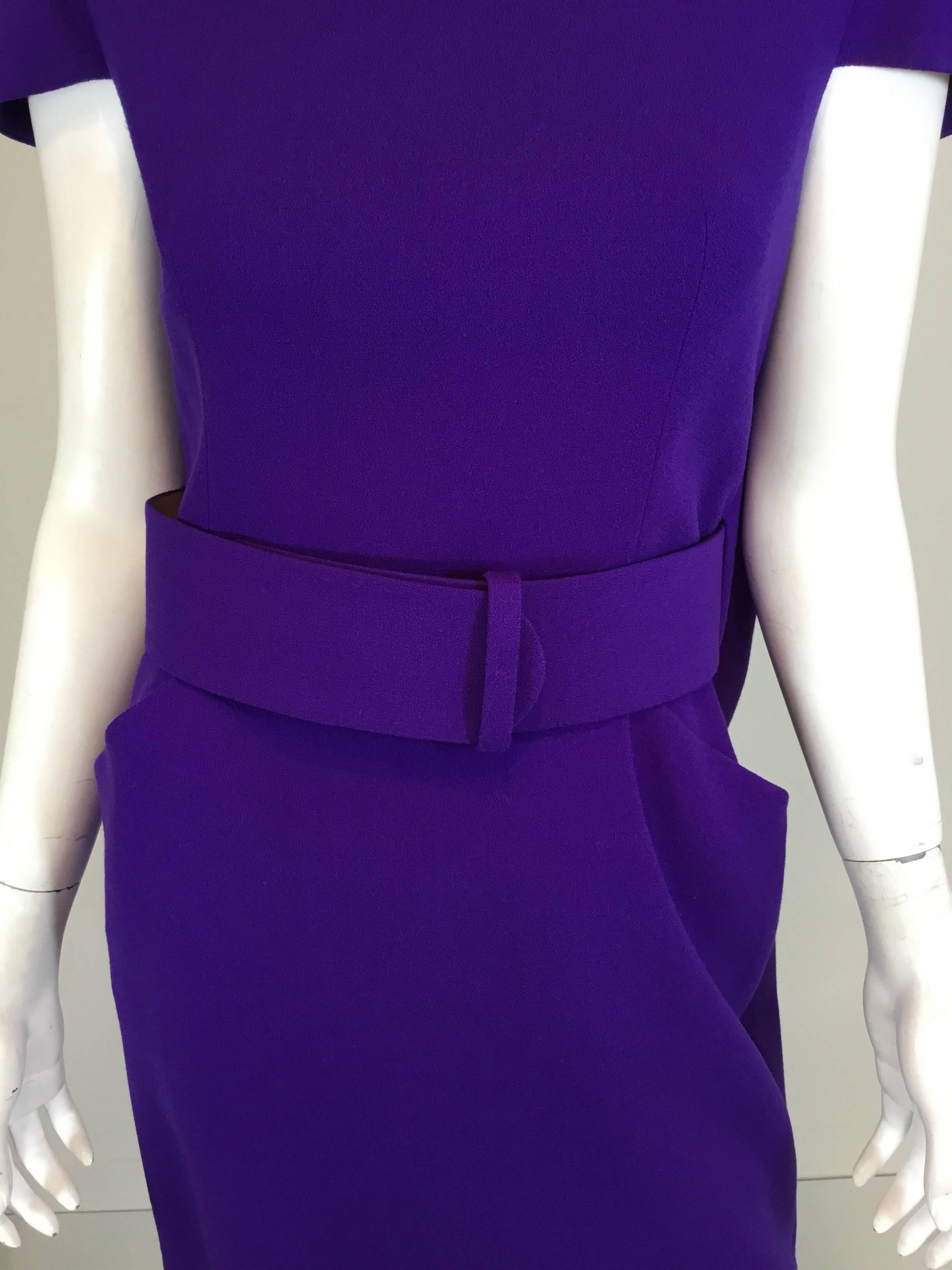 Alexander McQueen purple wool crepe dress features a belted waist (with a snap closure), pockets at the hips, capped sleeves, and a cape design at the back with a zipper fastening. Dress is fully lined, constructed with 100% wool and is made in