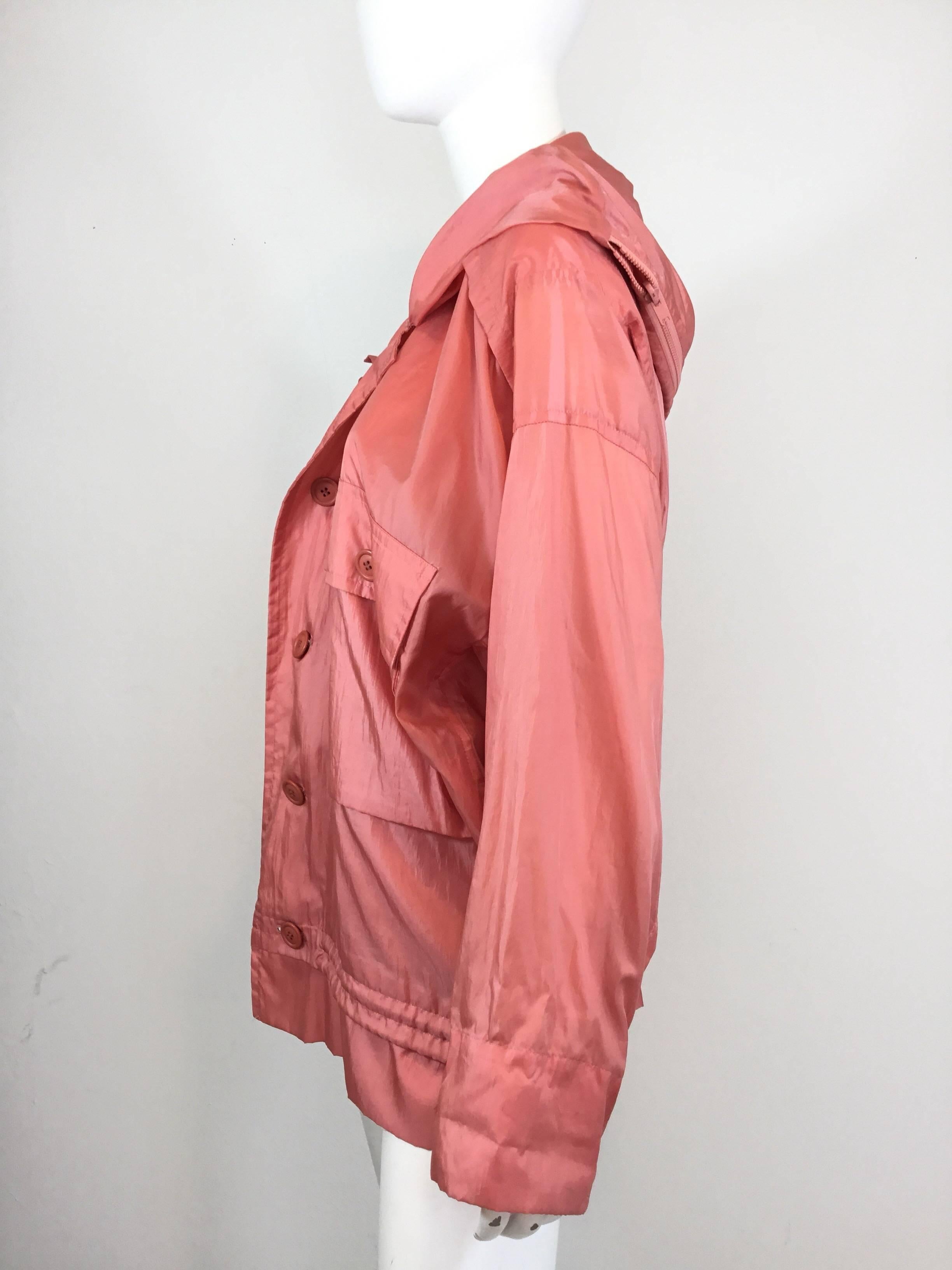Issey Miyake windbreaker jacket featured in a coral color. Jacket has a zippered front, large buttoned pockets at the bust, interior drawstring at the waist, and a hood in which has a zipper fastening to fold and store jacket inside. Labeled size S,