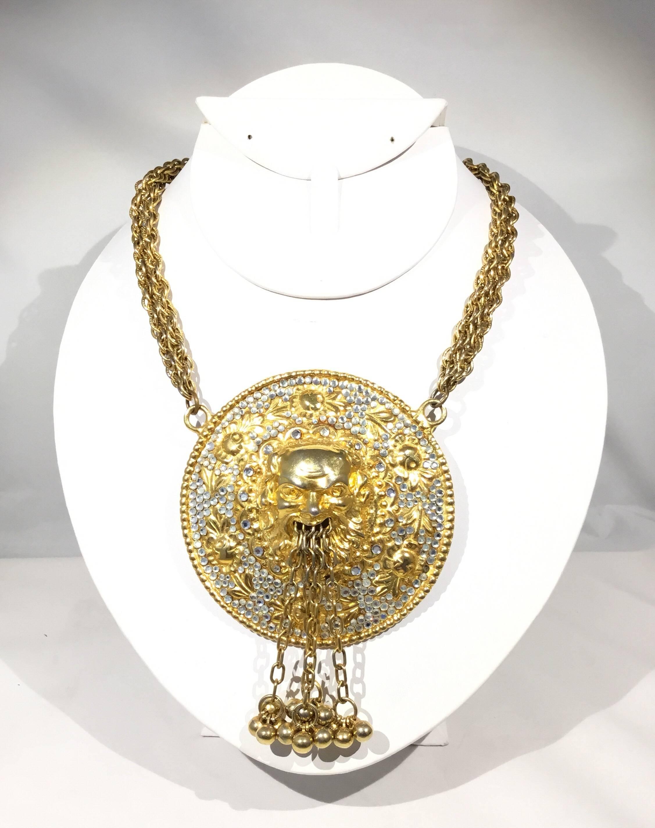 Die Judith Leiber Halskette Art to wear zeigt ein massives Medaillon, das Bachhus, den römischen Gott des Weins, darstellt. Aus dem Mund kommen baumelnde Perlenkugeln, die an den Rändern mit Strasssteinen besetzt sind. Doppelstrangige goldfarbene