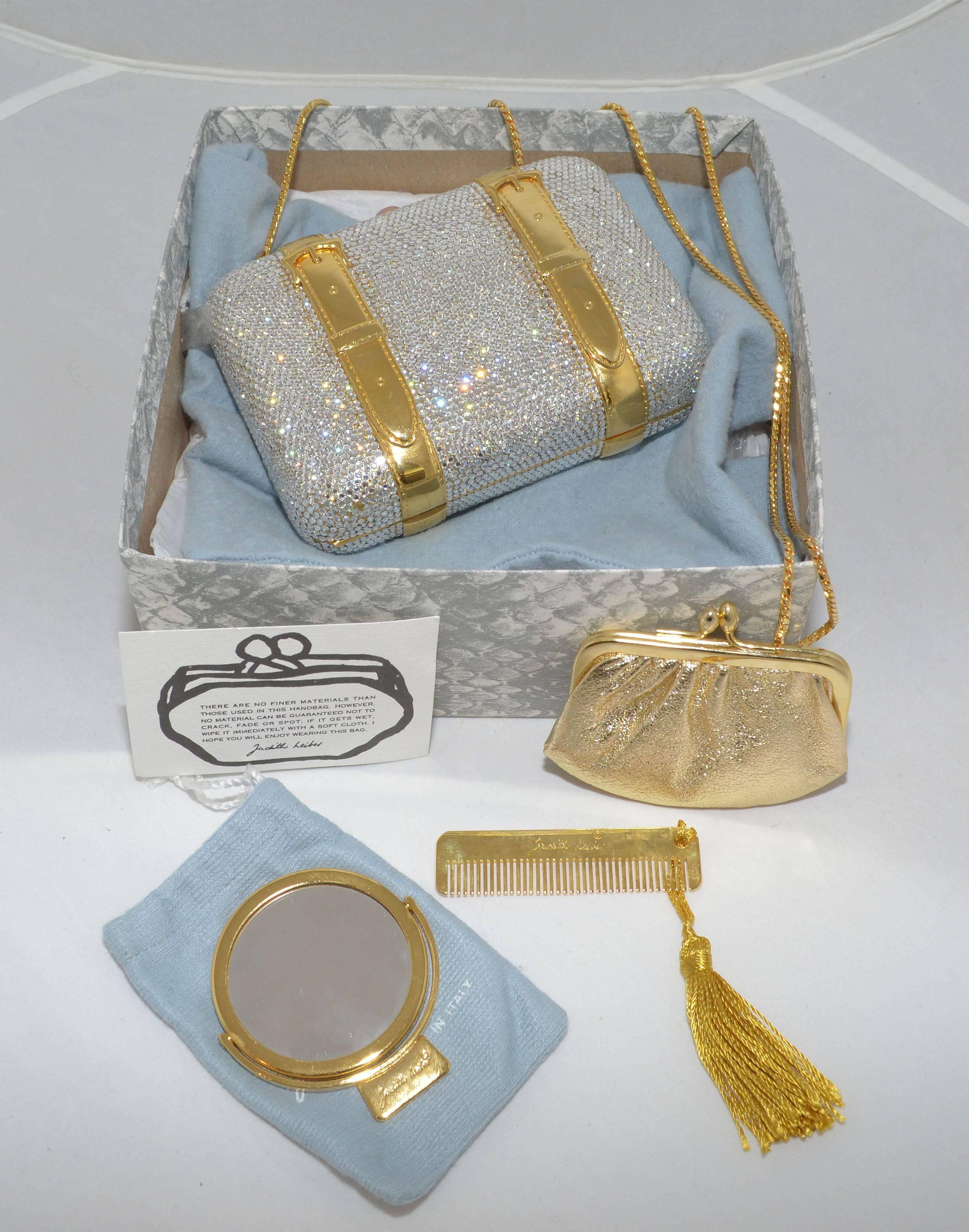 Stunning Judith Leiber clutch has a gold-tone decorative buckle strap detail, push top clasp closure, fully lined in a metallic gold leather, and includes: hand-held mirror, metal gold-tone comb, and a mini kiss lock coin pouch. Includes original